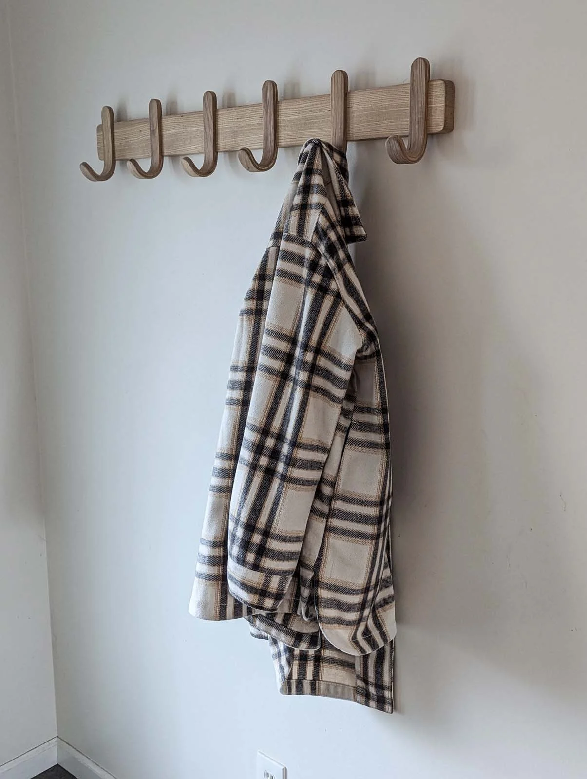 A wall-mounted wooden coat rack with seven hooks, holding a plaid shirt or robe in shades of beige, brown, and black.