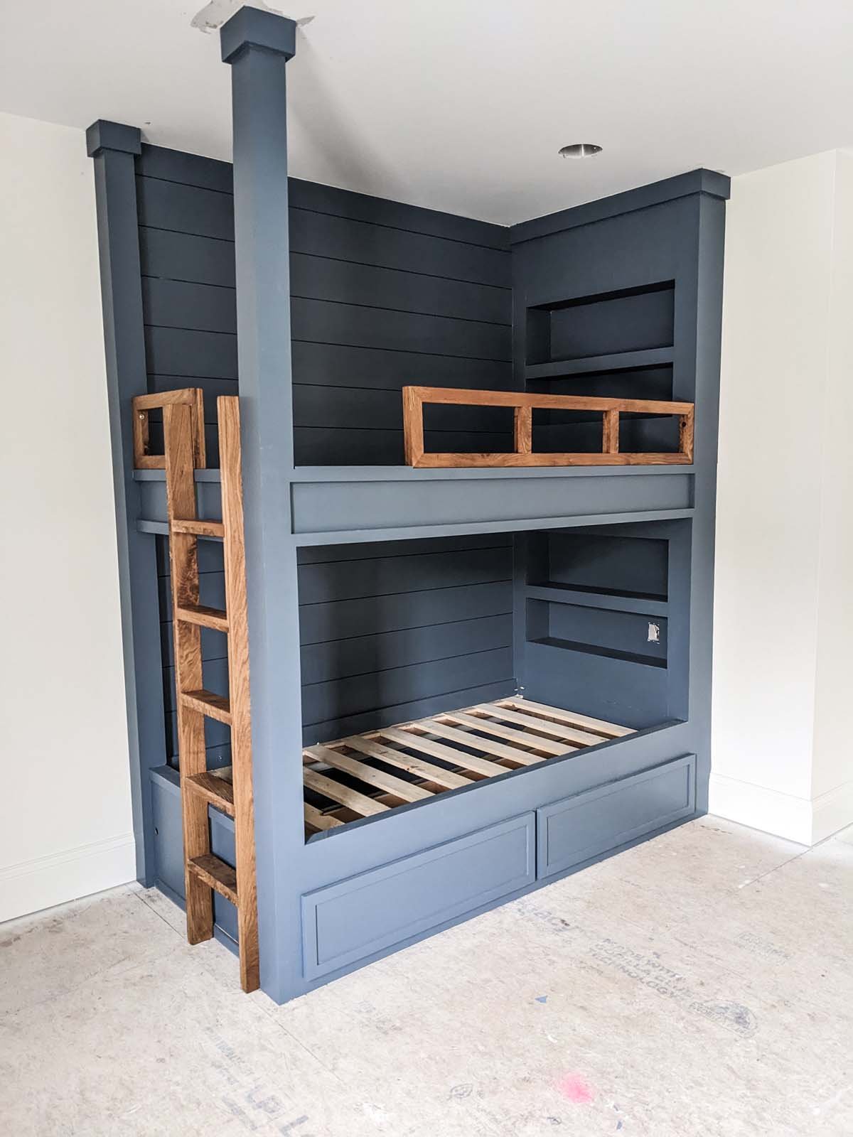 Custom built navy blue bunk bed with wooden ladder and safety rails, featuring a lower storage drawer and open shelving.
