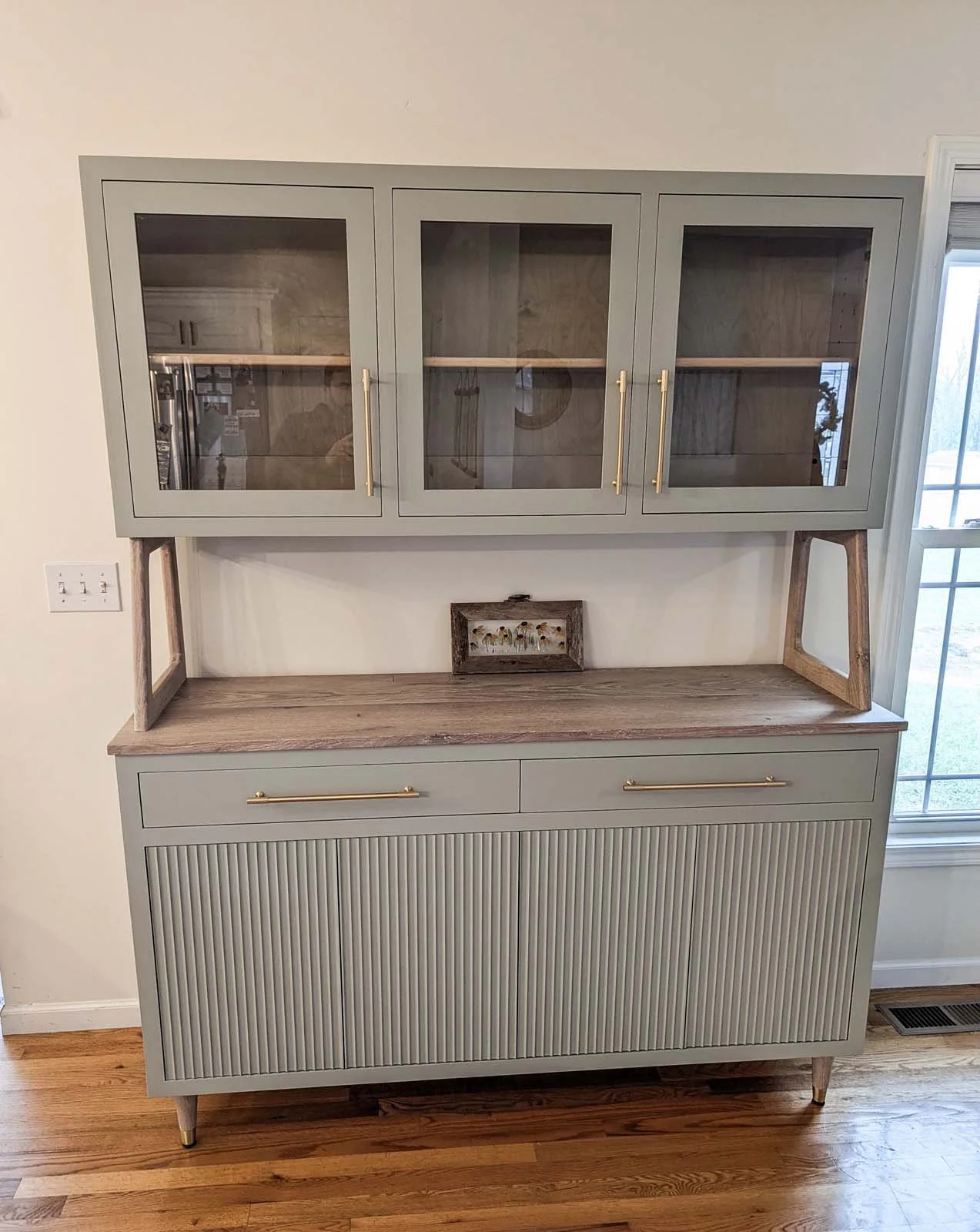 A light gray wooden hutch with glass-front upper cabinets and a solid lower cabinet with vertical grooved doors, styled with brass handles and trim, situated on a hardwood floor near a window.