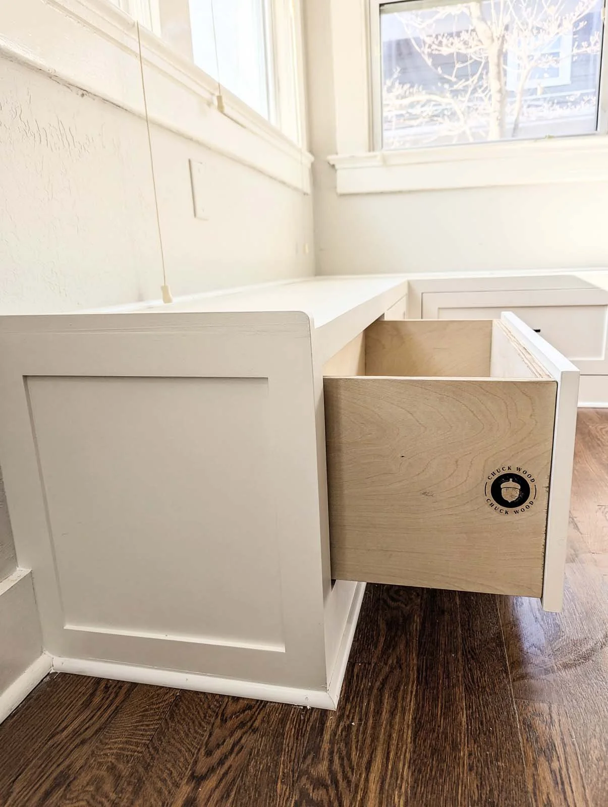 Empty white kitchen drawer partially pulled out from cabinetry, with a circular logo sticker on the front reading 'Chuck Wood,' surrounded by a black border. Wooden flooring and a window with a tree outside are visible.