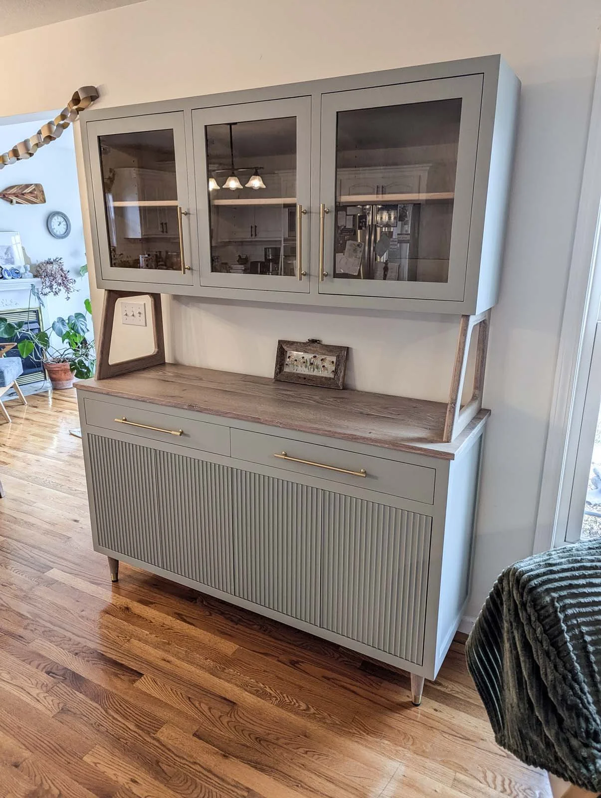 A light gray vintage sideboard with a wooden top and two brass handles, featuring glass-fronted upper cabinets, in a living room with hardwood floors and potted plants.