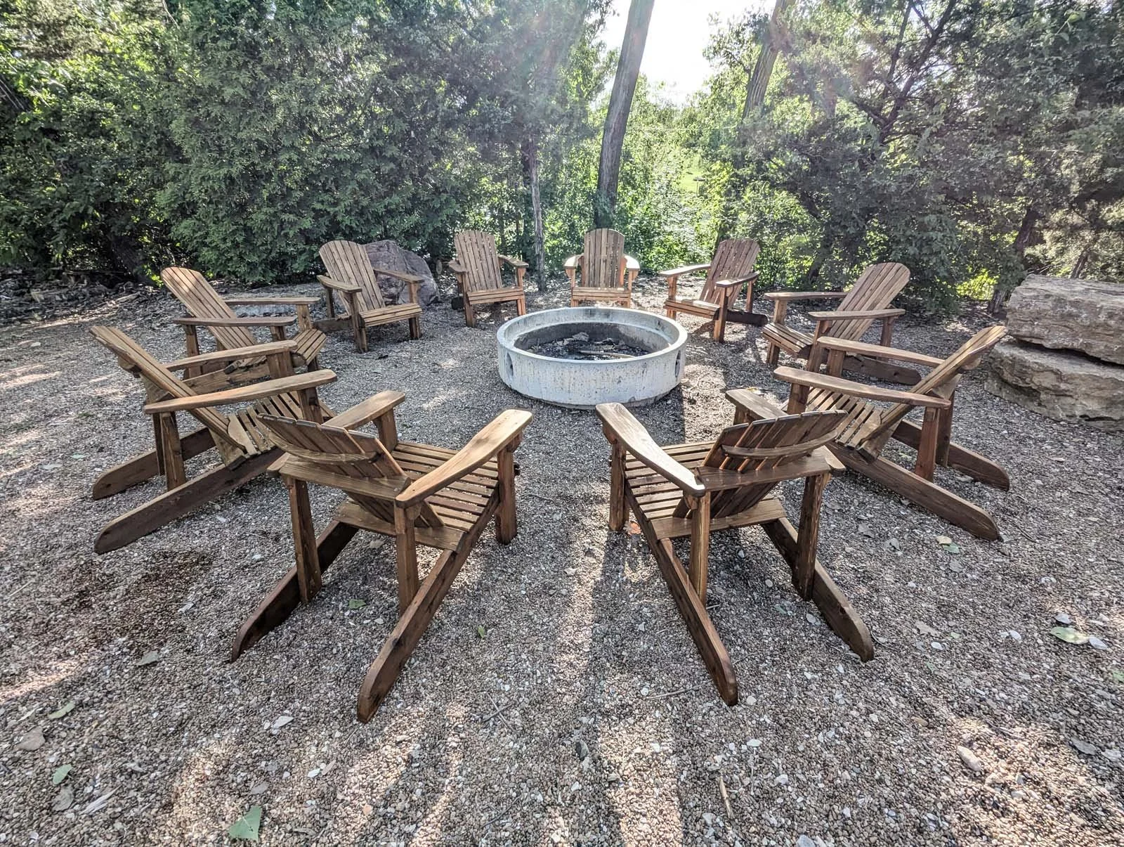 A circle of wooden Adirondack chairs surrounding a concrete fire pit in a wooded outdoor area.