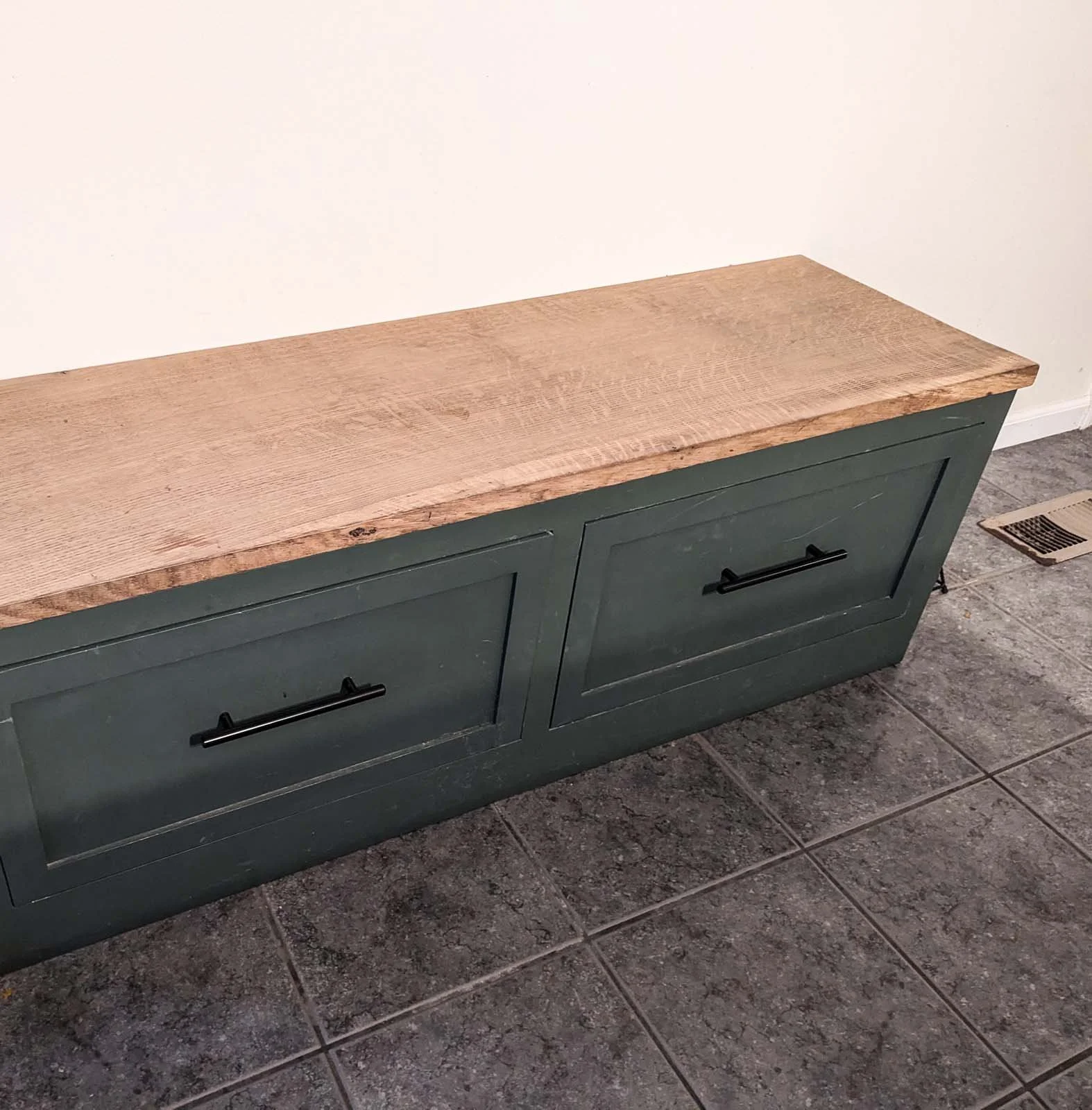 A wooden bench with a natural finish top and dark green painted base with two drawers, located next to a wall and on a tiled floor.