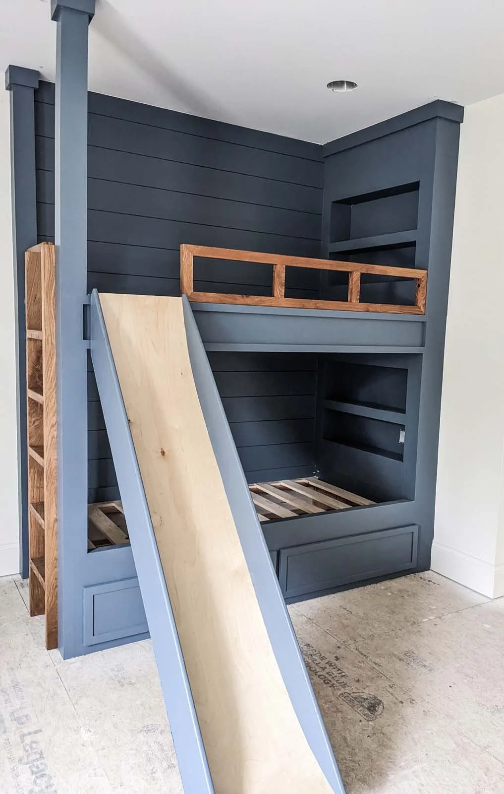 Construction of a custom built-in wooden structure with a slide, painted in blue and natural wood, in a room with beige tiled floor.