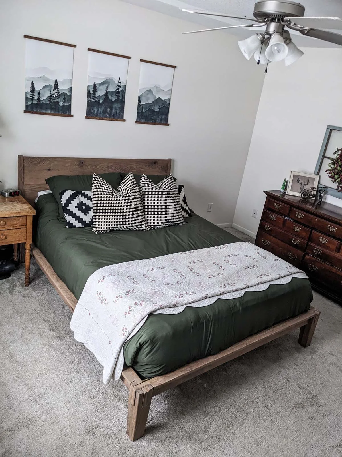 A bedroom with a wooden bed frame, green bedding, and black-and-white patterned pillows. Above the bed are three mountain landscape art prints. To the right of the bed is a wooden dresser with decorations including framed pictures and a wreath. The r