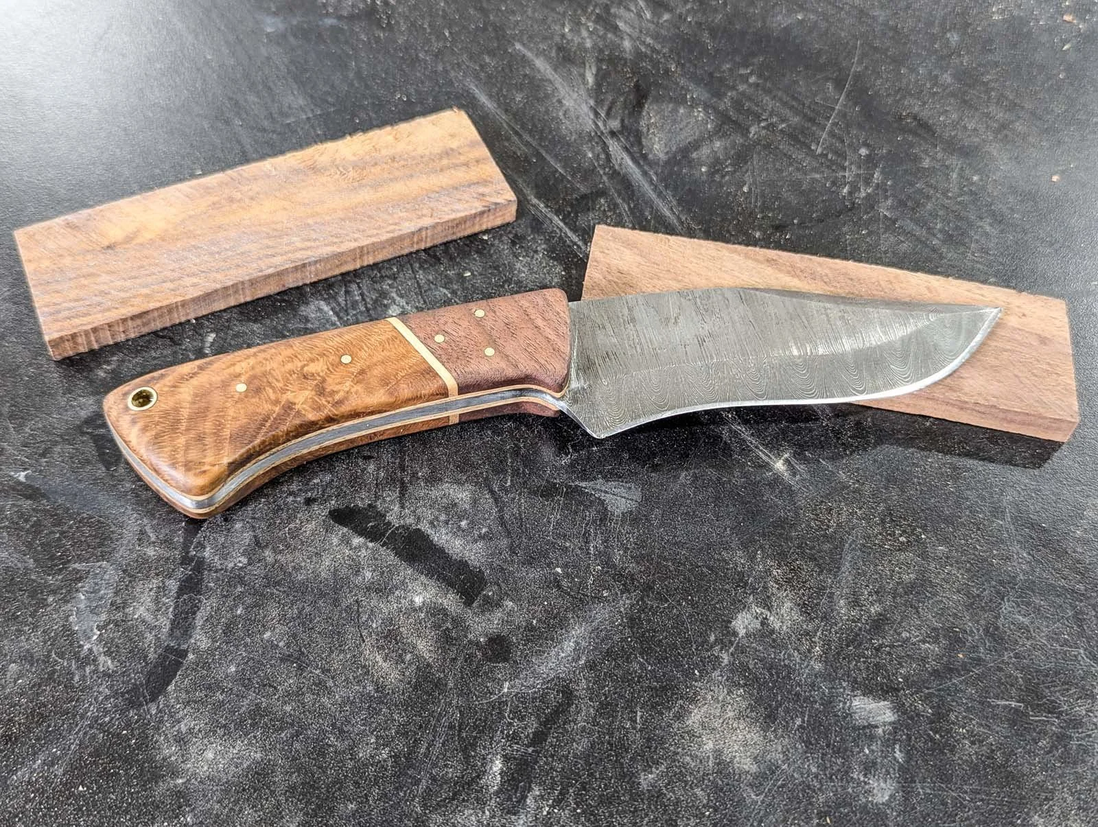 A woodworking knife with a wooden handle resting on a black workbench, with a piece of wood and a sharpening stone nearby.