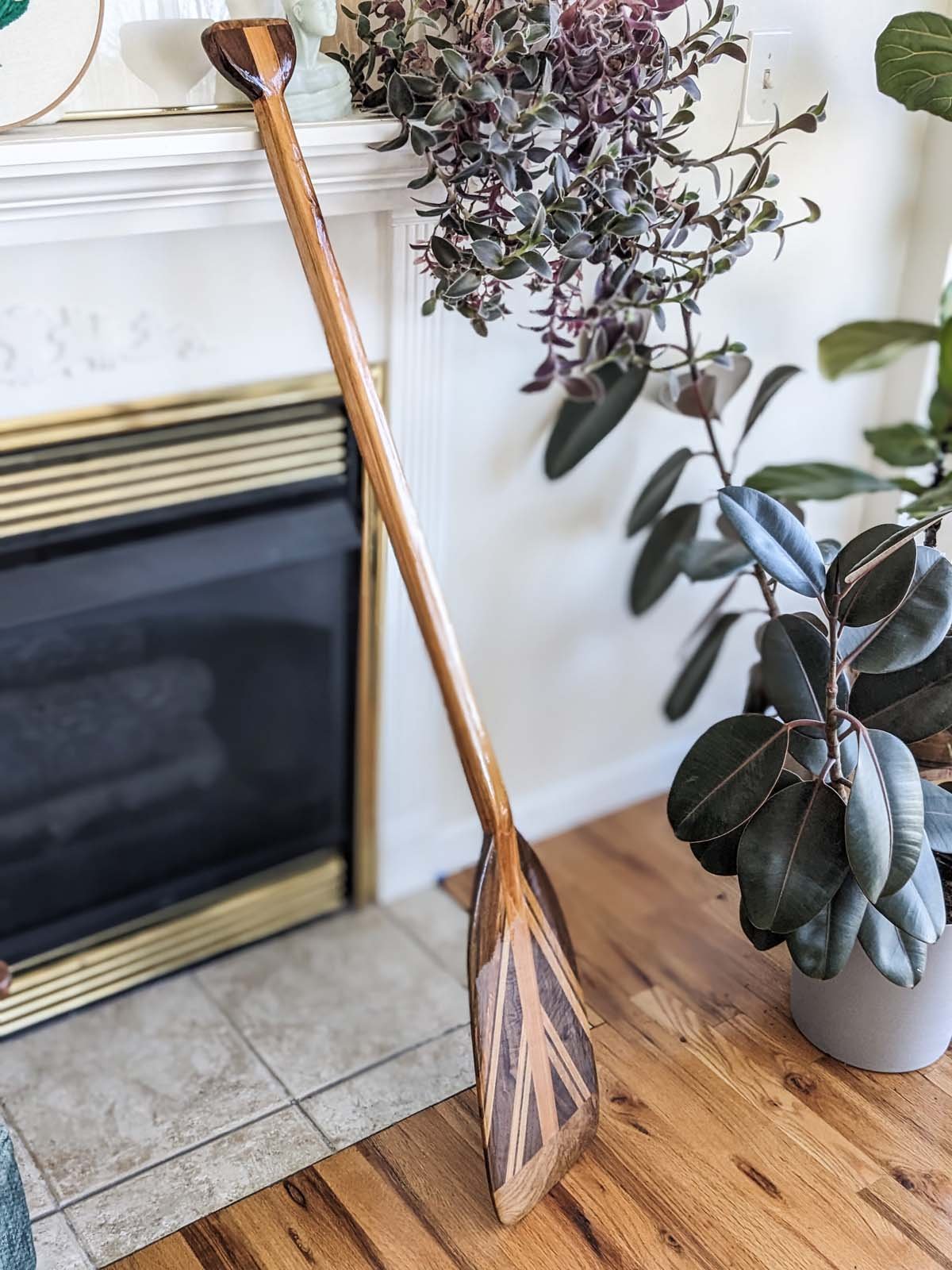 A wooden paddle leaning against a fireplace mantel surrounded by houseplants on a hardwood floor.