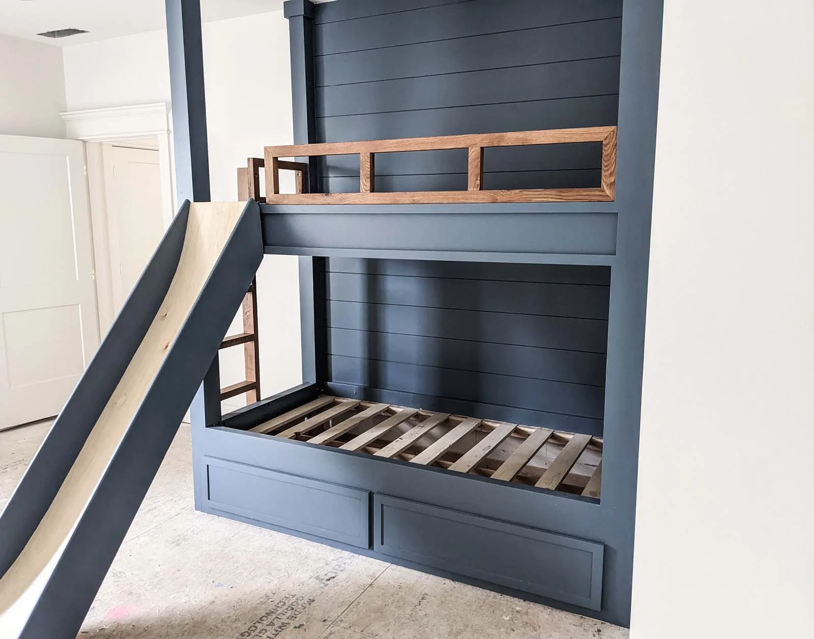 A built-in bunk bed frame with a slide and safety railing, painted dark blue with wood accents, inside a room with white walls and a hardwood floor.