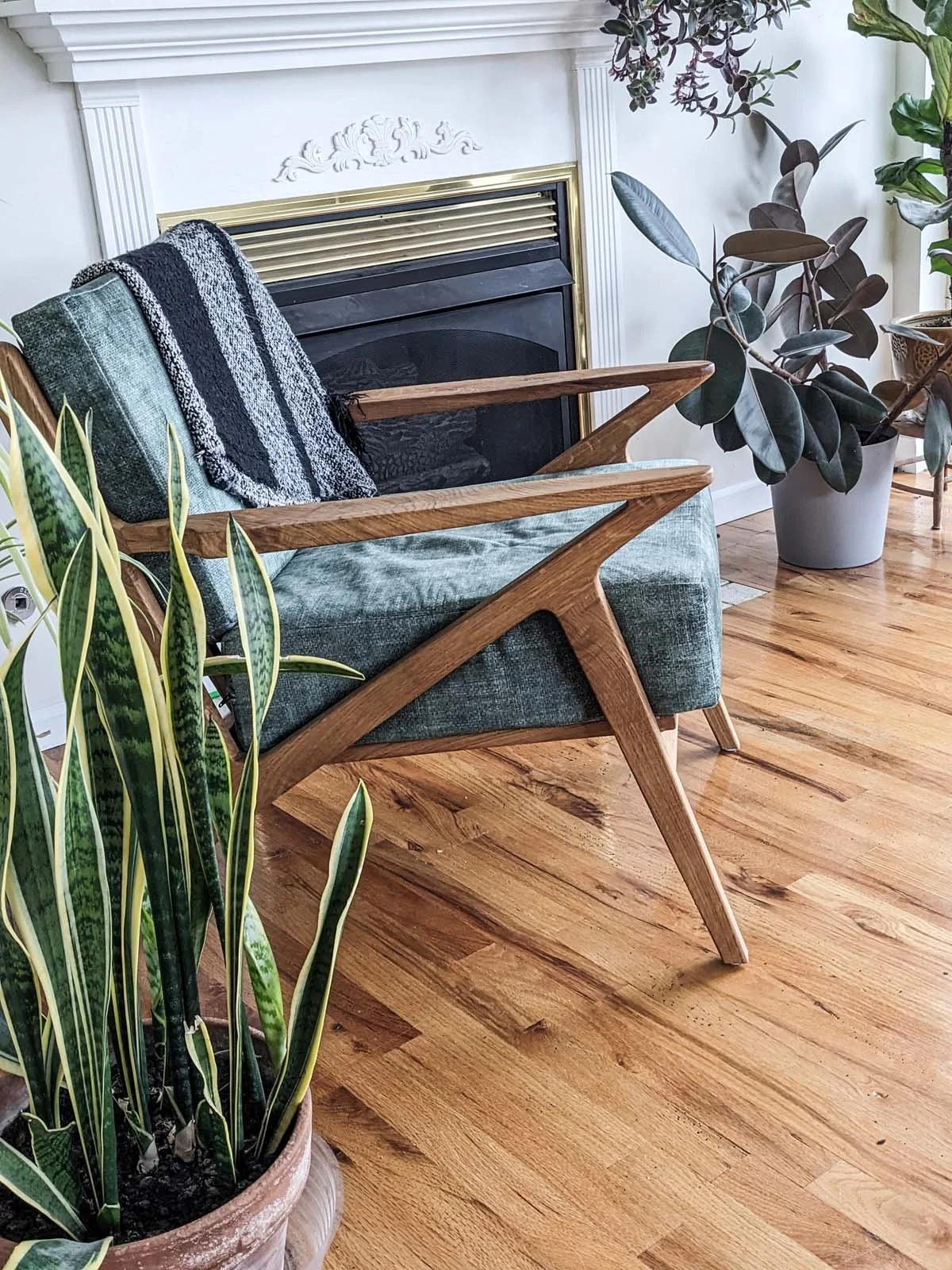 A mid-century modern armchair with grayish-green upholstery and wooden arms, positioned next to a fireplace with a decorative white mantel. Surrounding the chair are several houseplants, including a snake plant in the foreground and a larger plant wi