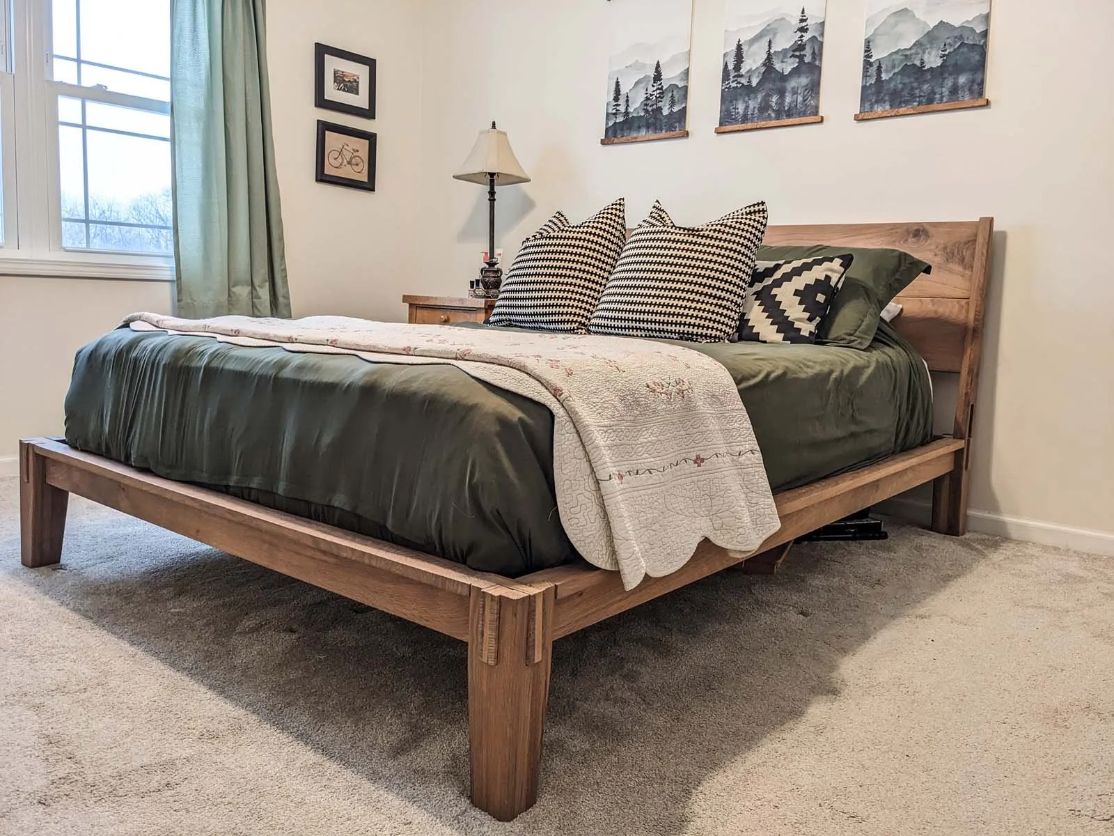 A bedroom with a wooden bed frame, green bedding, and patterned pillows, next to a wooden nightstand with a lamp, and wall art including mountain paintings and framed pictures of a bicycle and a landscape.
