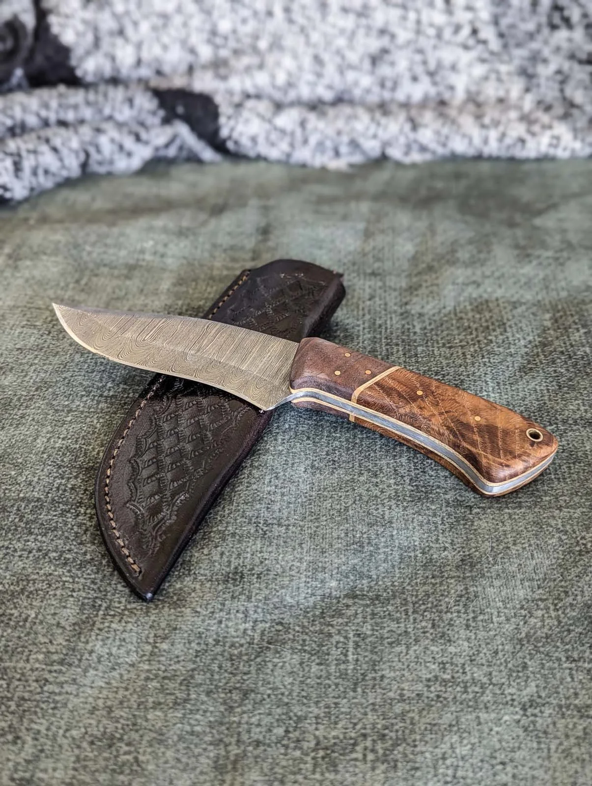 A handmade pocket knife with a patterned wooden handle and a Damascus steel blade, resting on a textured surface with a leather sheath nearby.