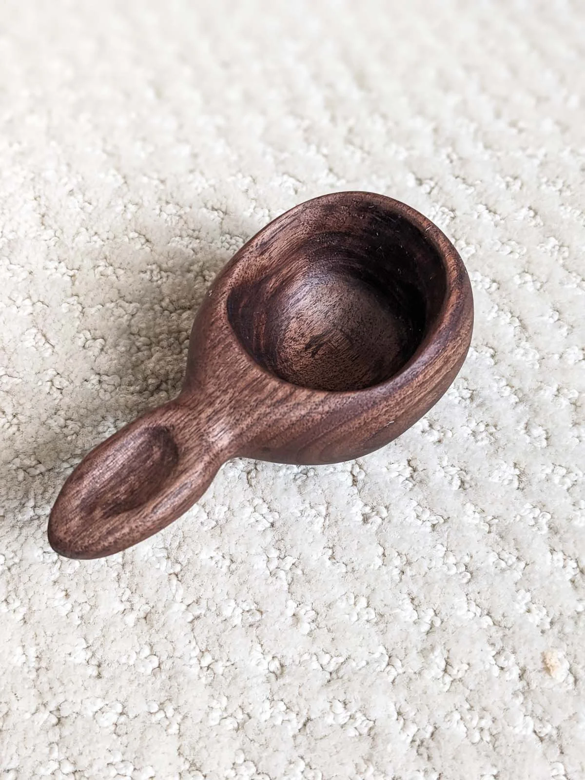 A small, dark wooden scoop with a handle, resting on a textured, light-colored surface.