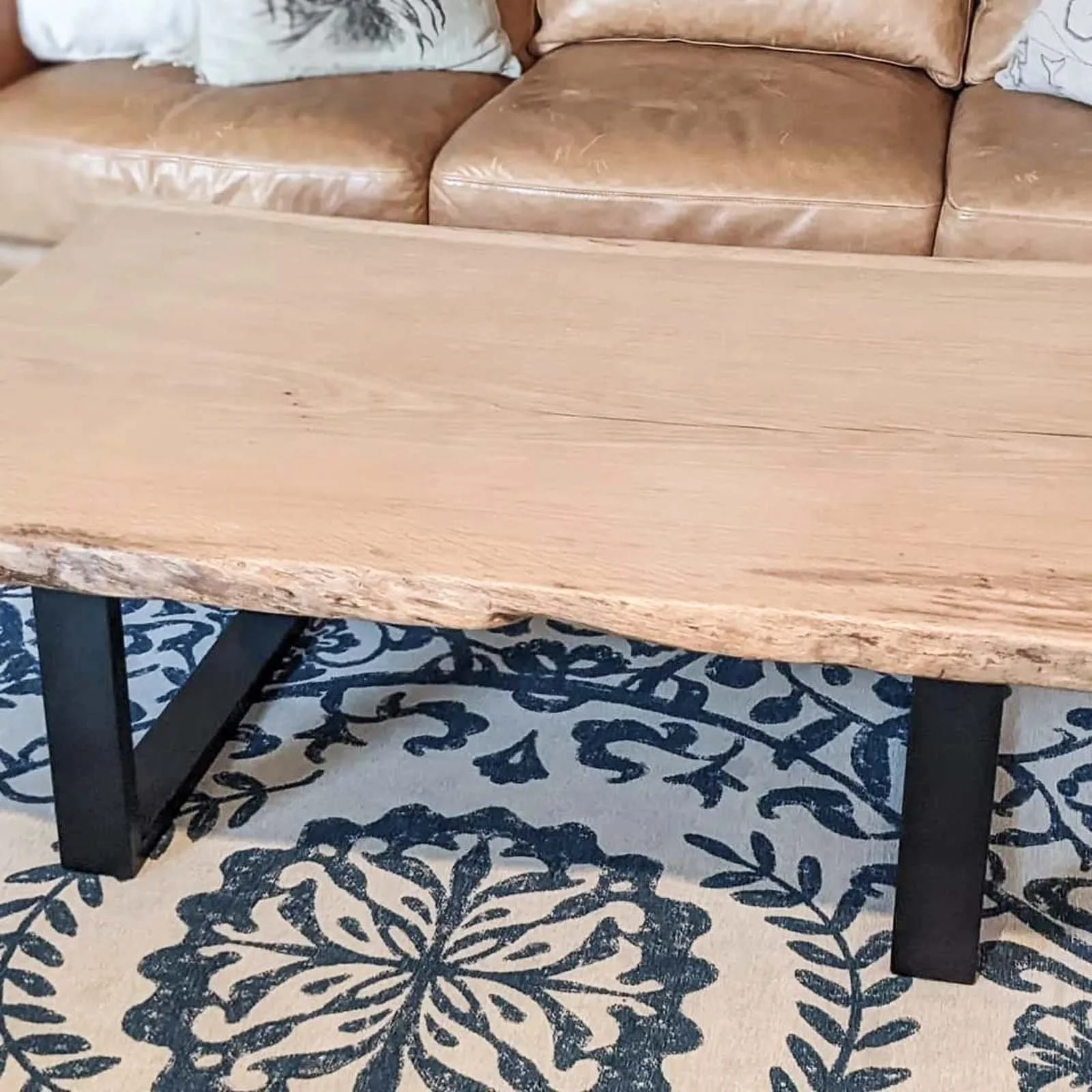 A close-up of a wooden coffee table with black legs, placed on a blue and white patterned rug, in front of a tan leather sofa with pillows.
