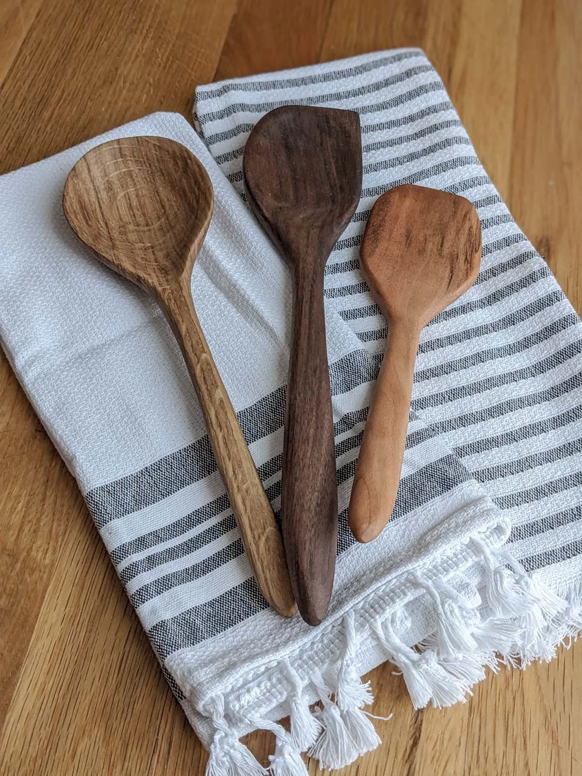 Three wooden spoons of different sizes and colors resting on a black and white striped cloth on a wooden surface.