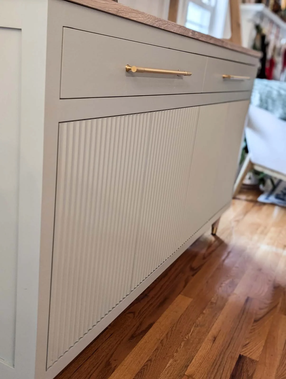 A beige sideboard with gold handles, featuring a ribbed design on the lower section and a smooth upper section with drawers, standing on a wooden floor in a well-lit room.