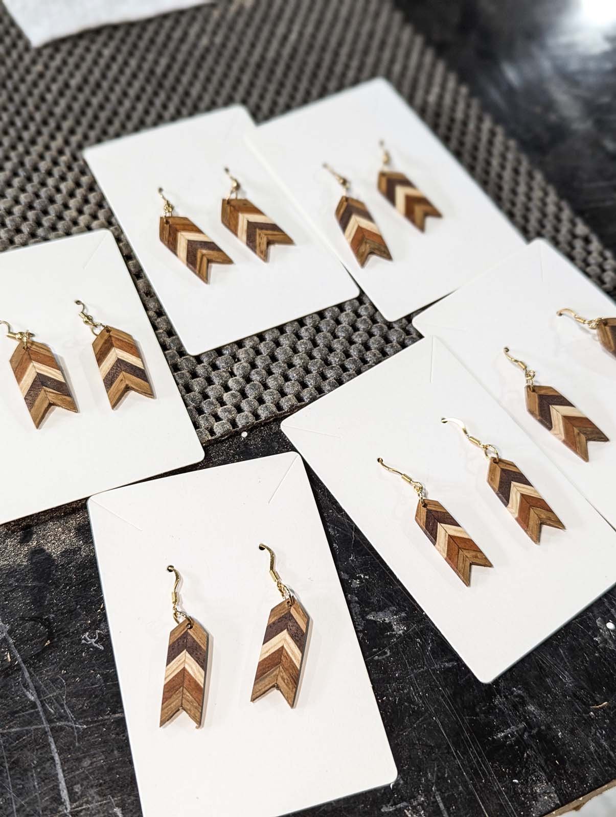 Multiple pairs of wooden geometric earrings with chevron pattern, displayed on white cards, on a black textured surface.
