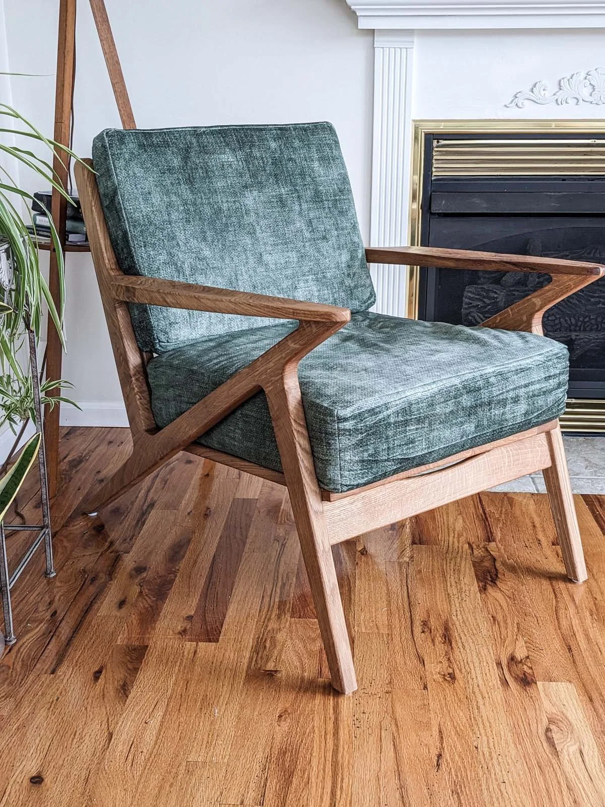 A mid-century modern armchair with a wooden frame and teal velvet cushions, placed on a hardwood floor in front of a fireplace.
