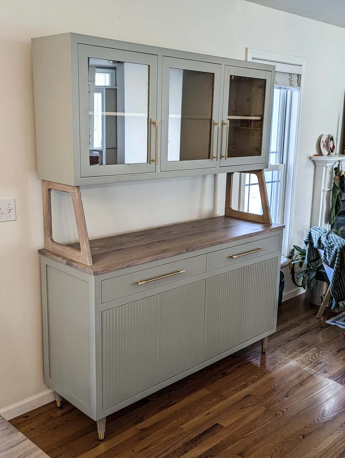 A vintage-style cabinet with glass doors on top, mounted on a wooden stand attached to a lower base with storage drawers, in a room with hardwood flooring.