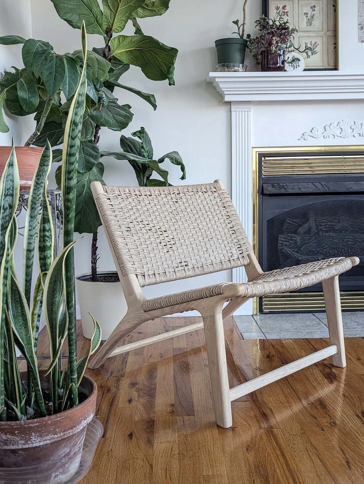 Living room corner with green plants, a small wooden chair with woven seat, and a fireplace with a marble mantel.