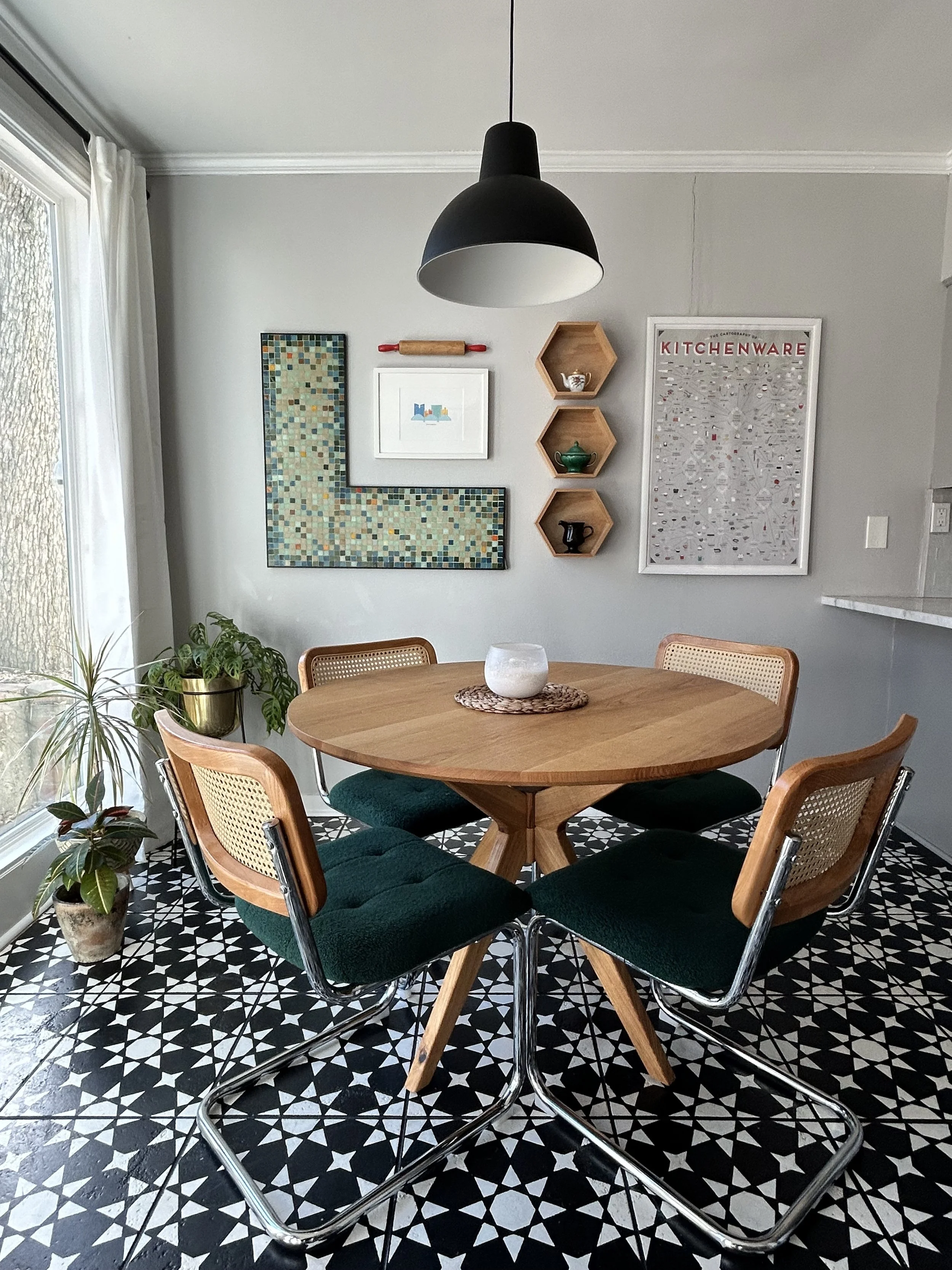 A dining area with a round wooden mid century modern table, four chairs with green cushions, a black hanging pendant light, potted plants near a window, and wall decor including a mosaic art piece, small hexagonal shelves, and a kitchenware poster.