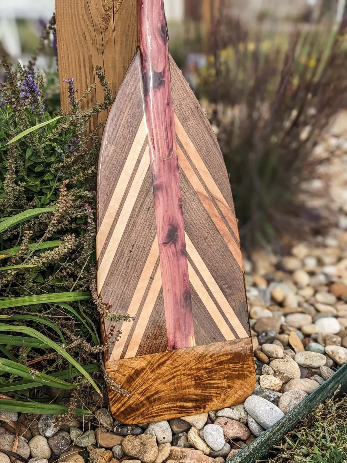 A decorative wooden garden ornament with a triangular shape, featuring various wood patterns and a pinkish streak in the center, placed on a bed of small, multicolored stones next to some green plants and purple flowers.