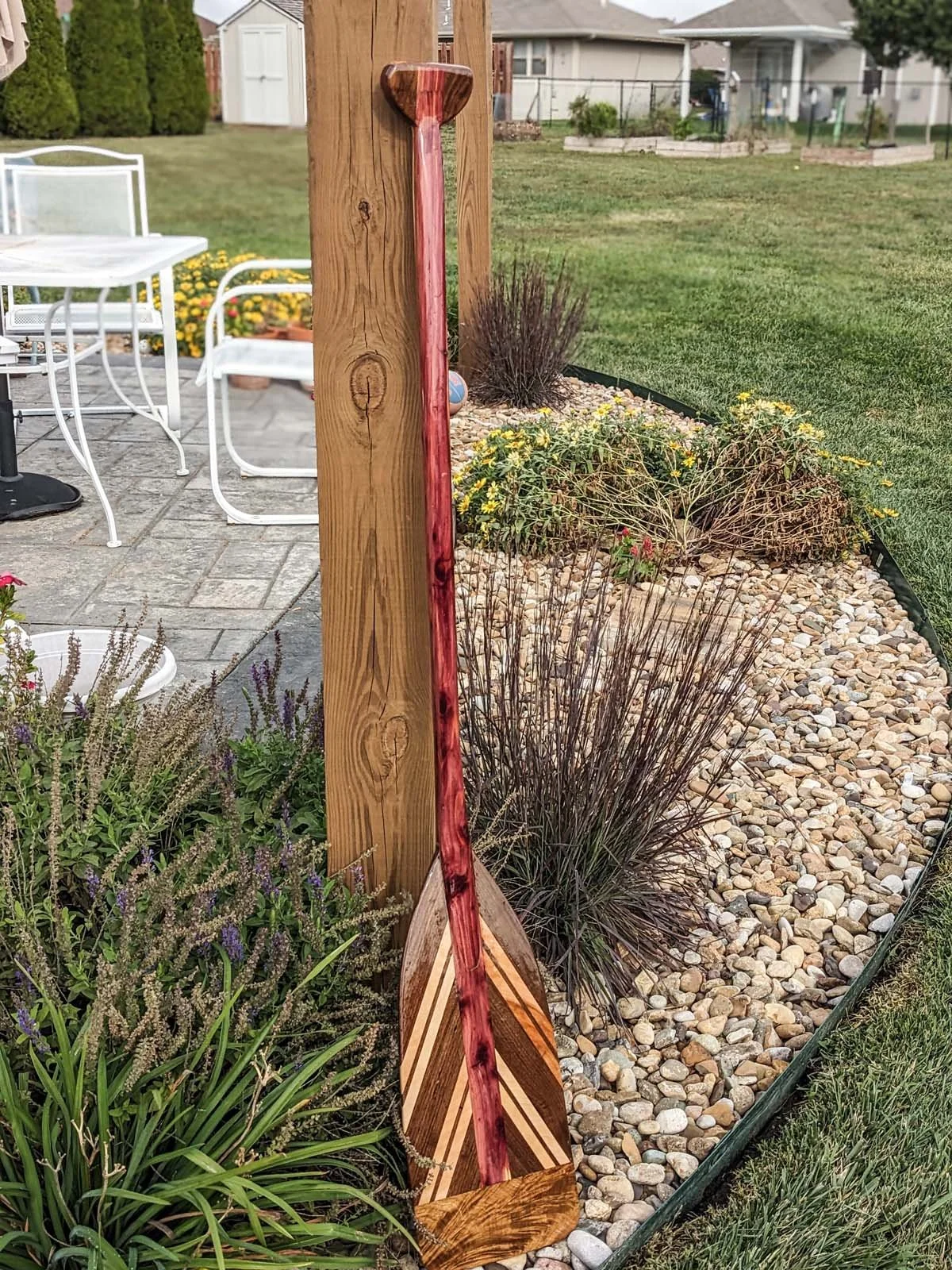 A wooden garden stake with a decorative top, placed in a flower bed with rocks, plants, and bushes, near patio furniture in a backyard.