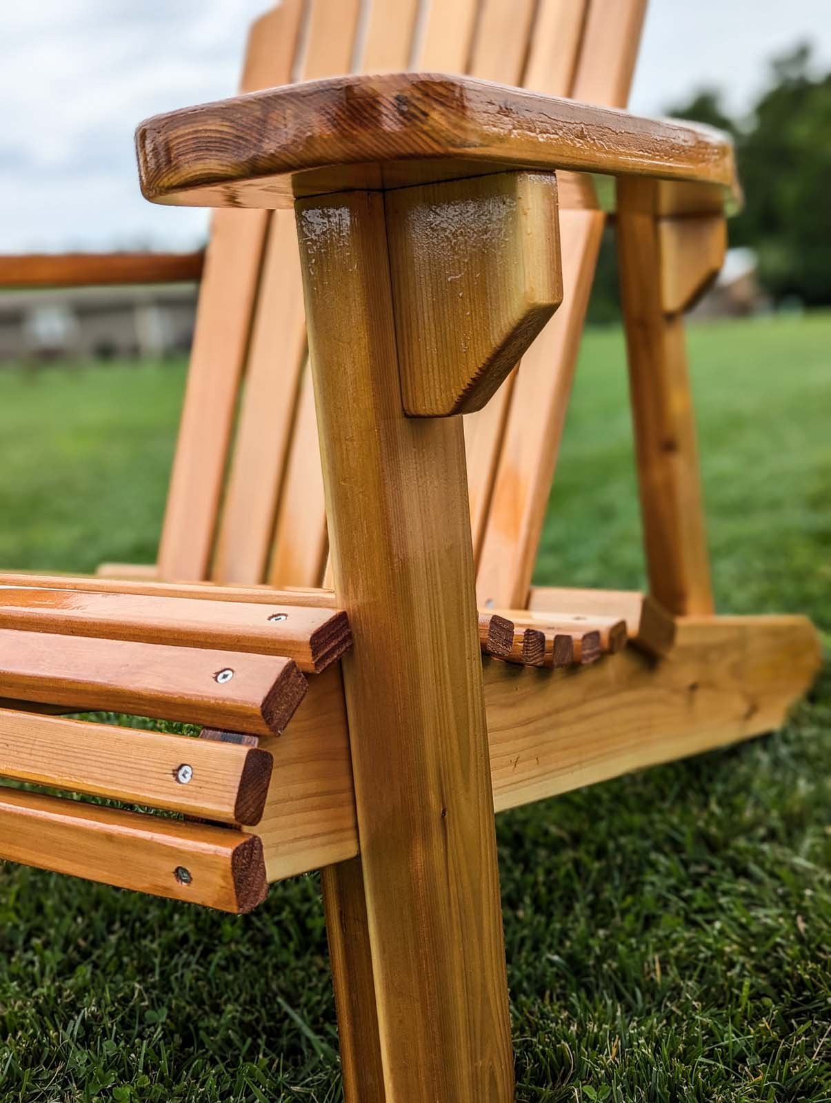 Close-up of a wooden outdoor bench with slatted backrest and armrest, situated on a grassy lawn with trees and a cloudy sky in the background.