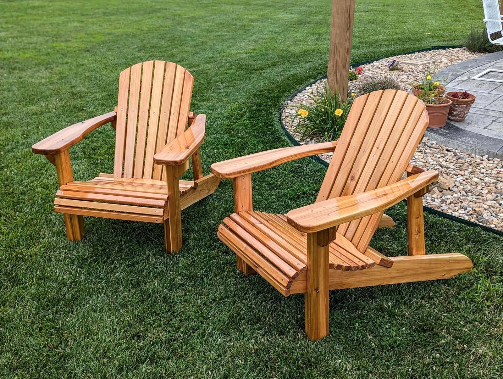 Two wooden Adirondack chairs on a green lawn near flower beds with pots and a stone patio.