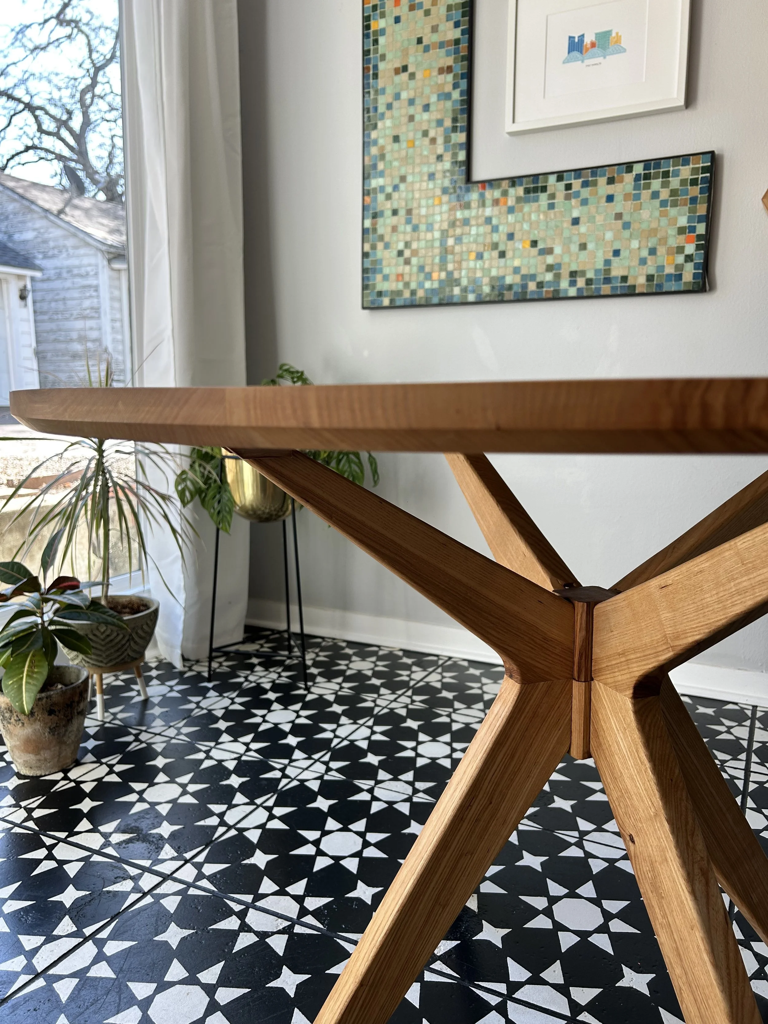 Interior view of a room with a wooden table, patterned black and white floor tiles, potted plants, and framed artwork on the wall.