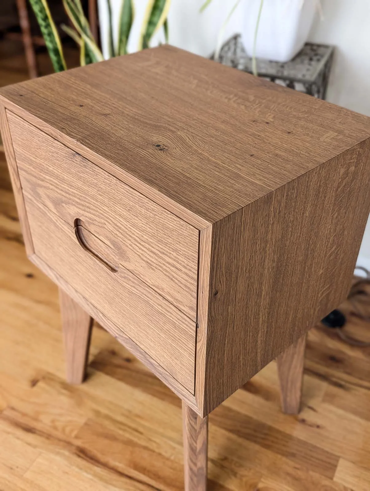 A wooden nightstand with a single drawer and a curved handle, standing on four angled legs on a wooden floor.