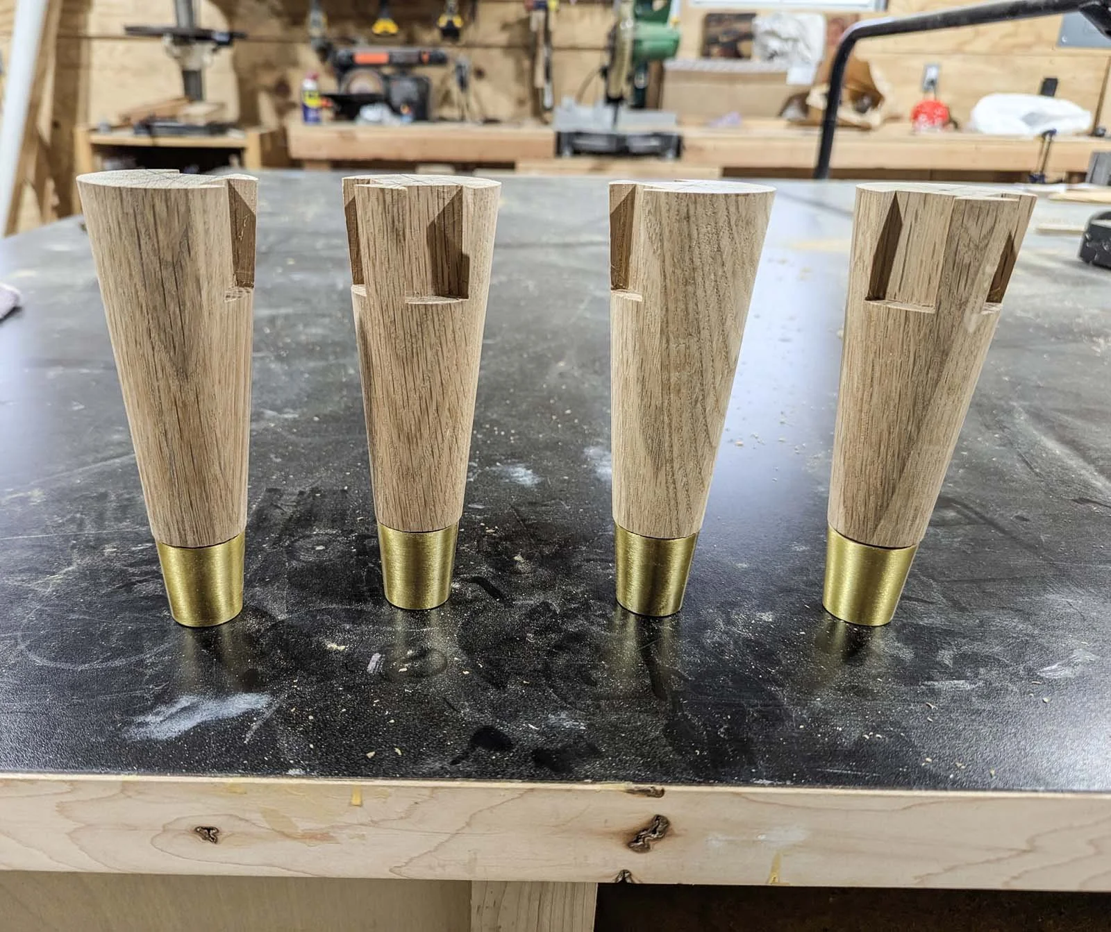 Four wooden furniture legs with gold-colored metal tips on a workbench in a woodworking shop.