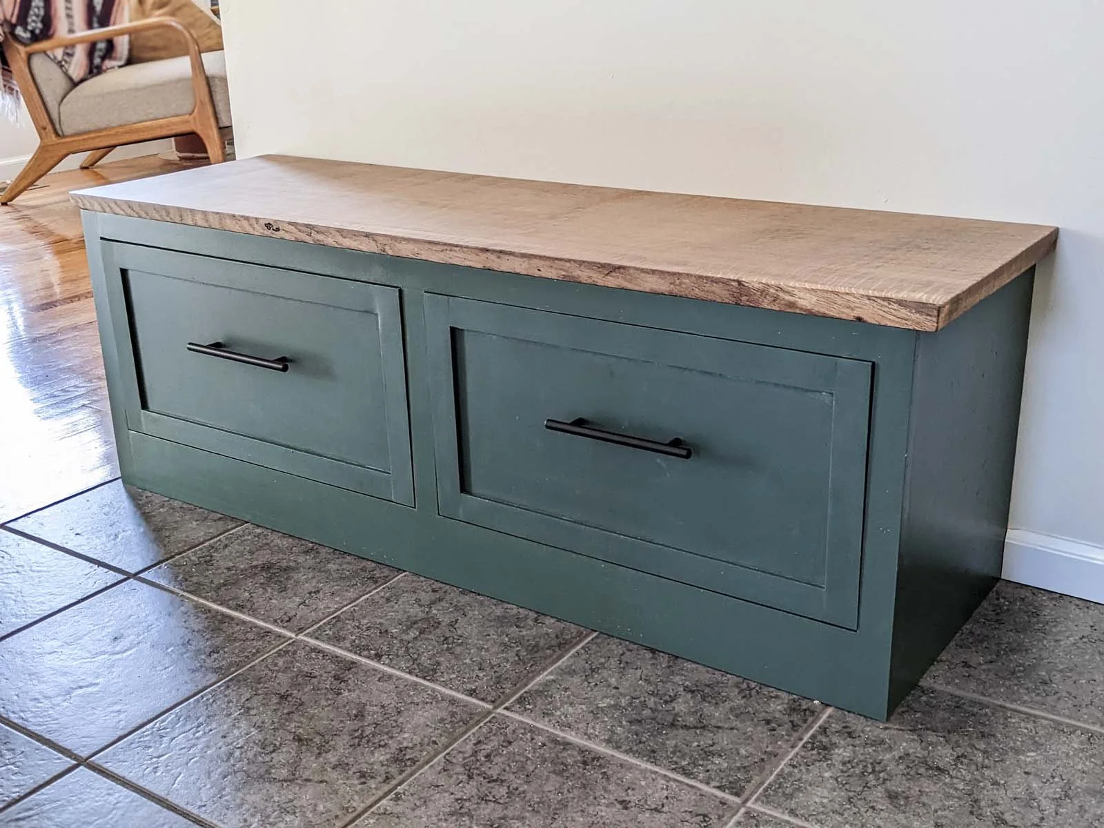 A wooden bench with a painted green base and a natural wood top, placed against a wall in a room with tiled floor and a chair in the background.