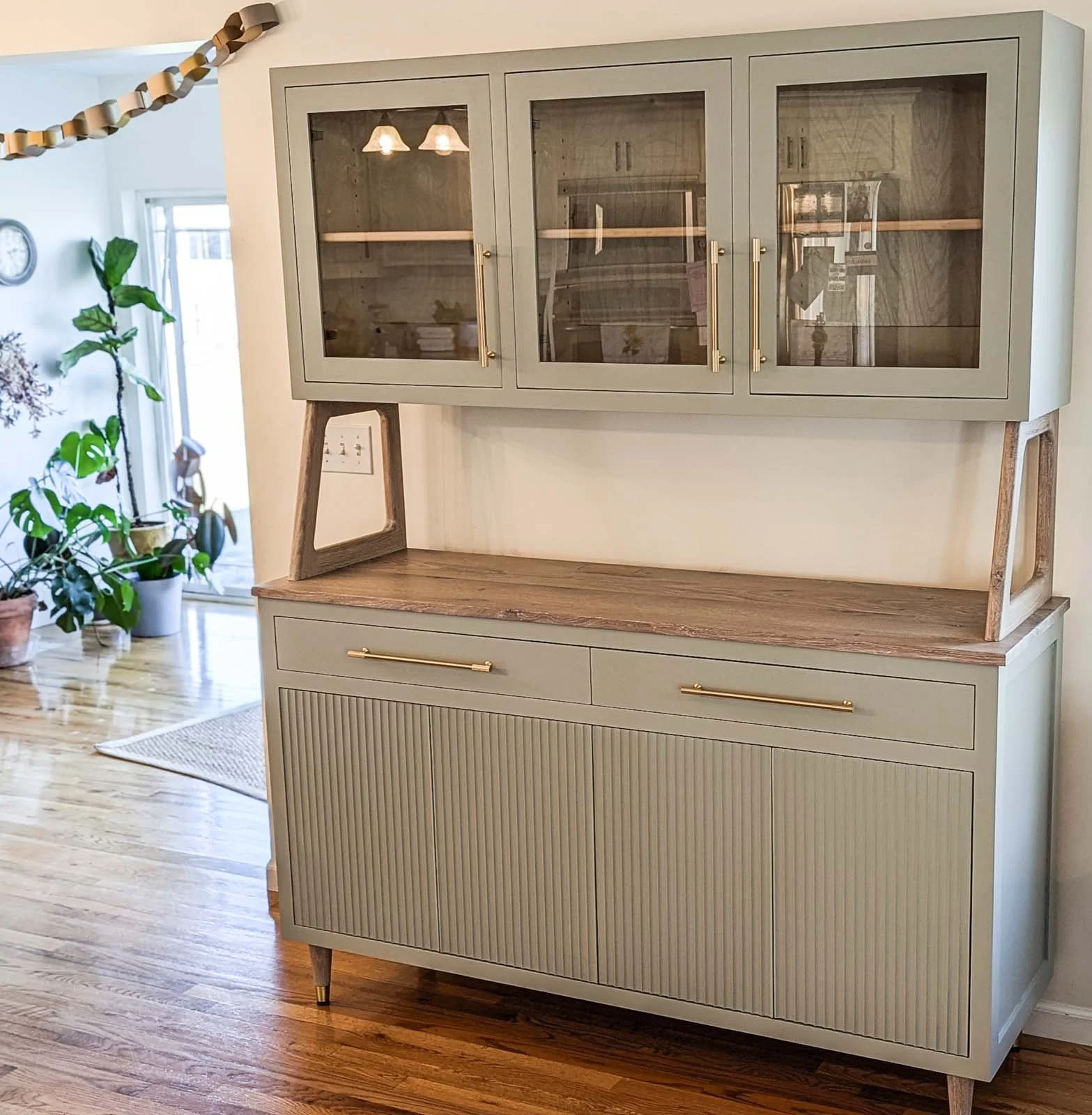 A two-tone vintage sideboard with a light green lower cabinet and glass-fronted upper cabinet in a room with hardwood floors and potted plants.