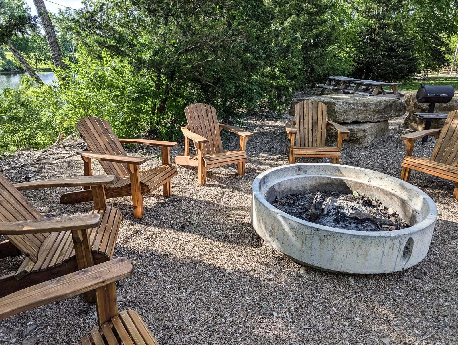 A circle of wooden Adirondack chairs surrounds a concrete fire pit with ashes inside, set outdoors on gravel near trees and a body of water.