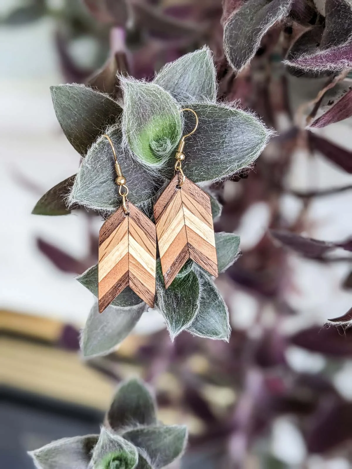 Wooden arrow-shaped earrings hanging on a leafy plant with fuzzy, gray-green leaves.
