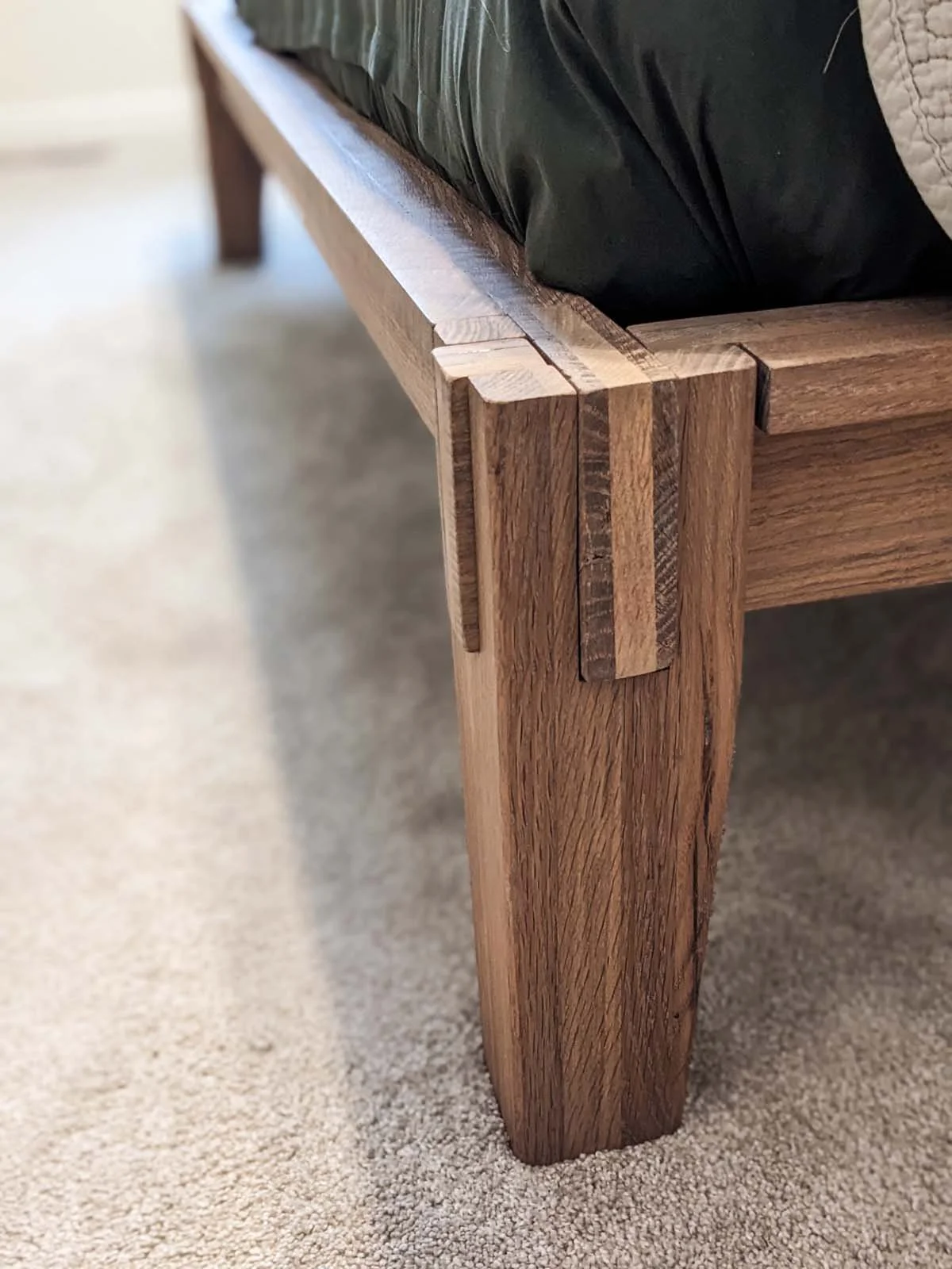 Close-up of a wooden bed frame corner with a dovetail joint, on a beige carpet.