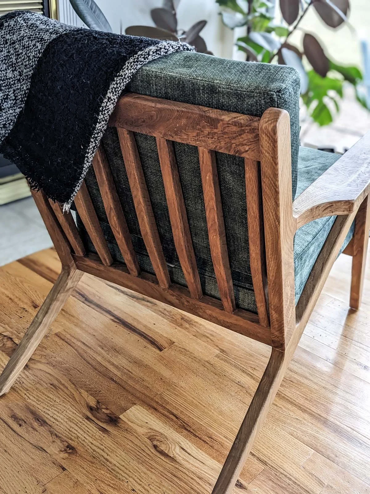 A wooden-framed armchair with dark green fabric cushions, placed on a wooden floor, with a black textured throw blanket draped over the back.