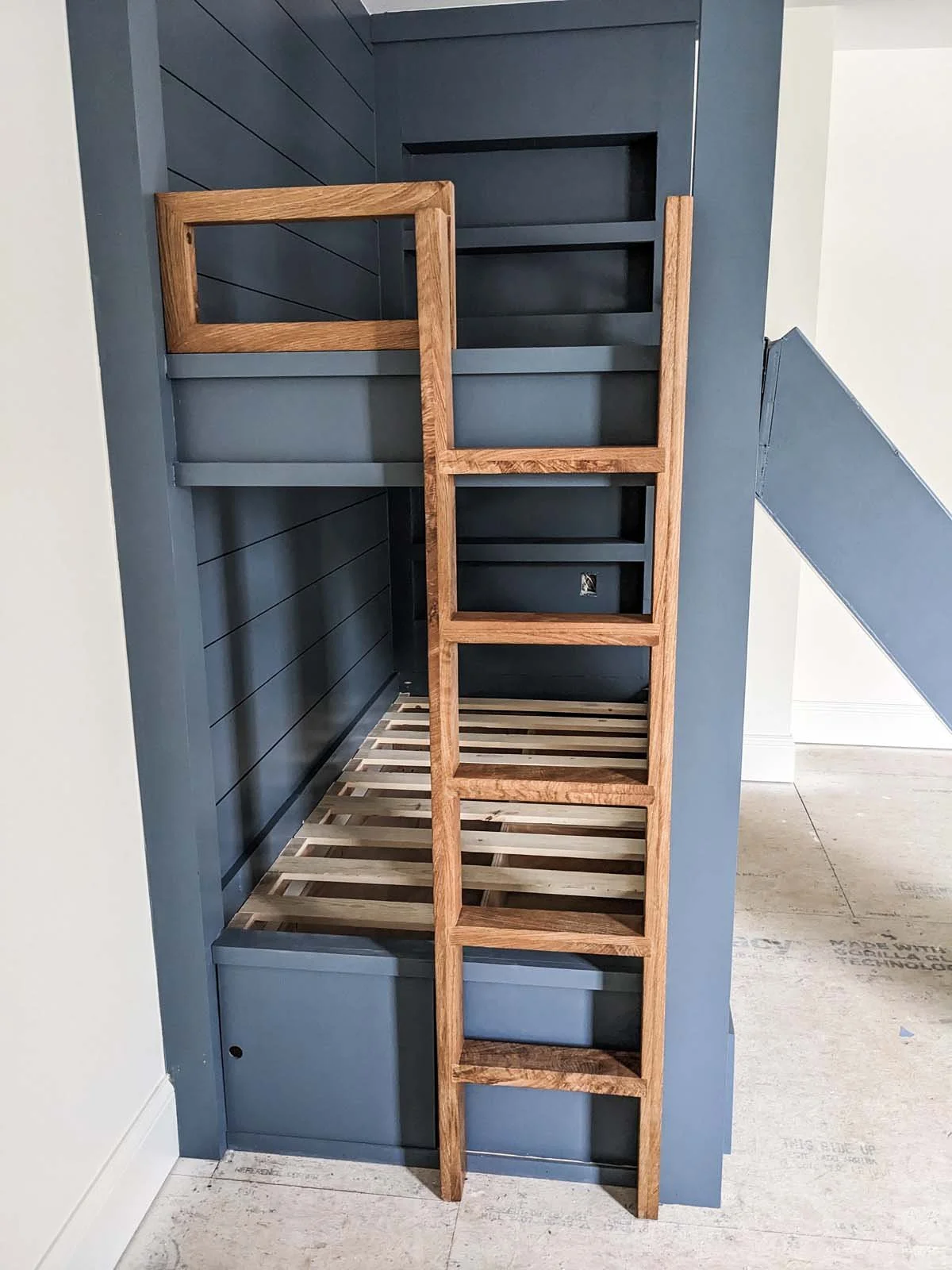 Unfinished built-in bunk bed with wooden ladder, painted dark blue background, and slatted base.