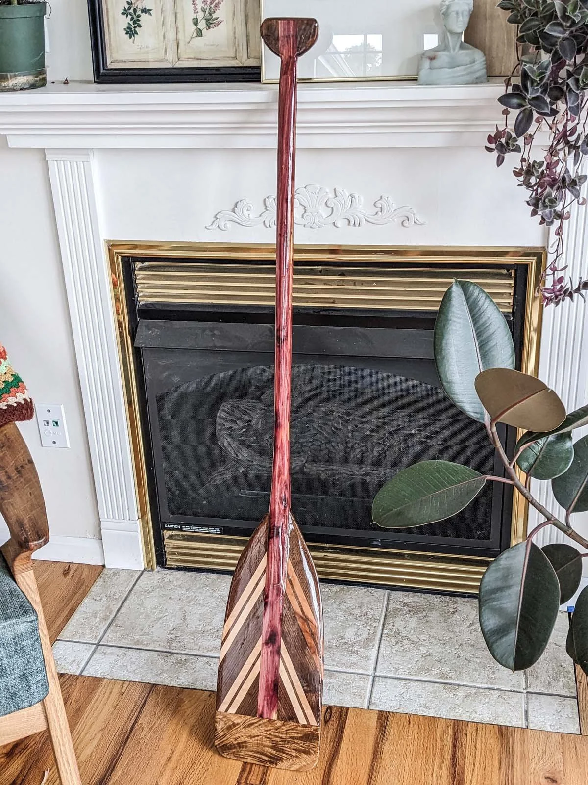 A decorative wooden paddle with a geometric pattern on the blade, leaning against a white fireplace with ornate molding and black and gold accents.