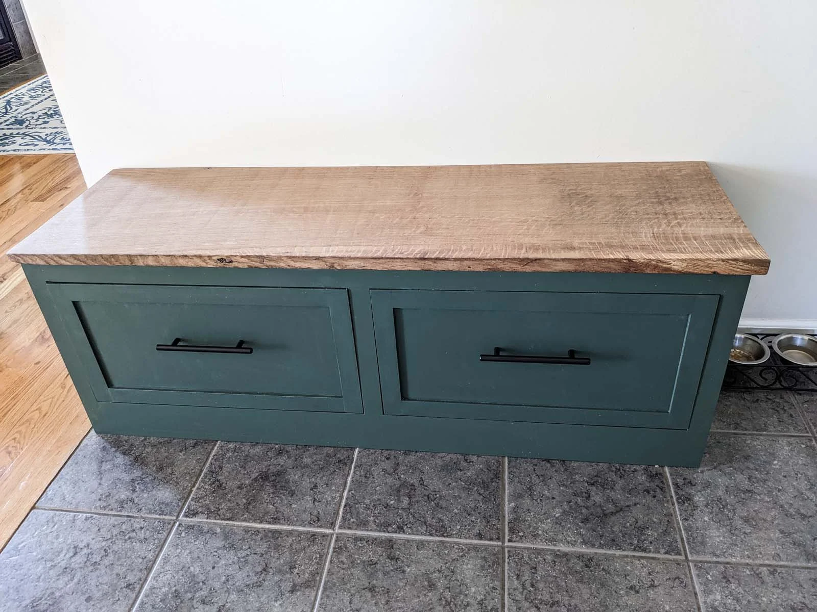 A wooden bench with a natural finish top and teal painted base, located on a tiled floor in a home interior.