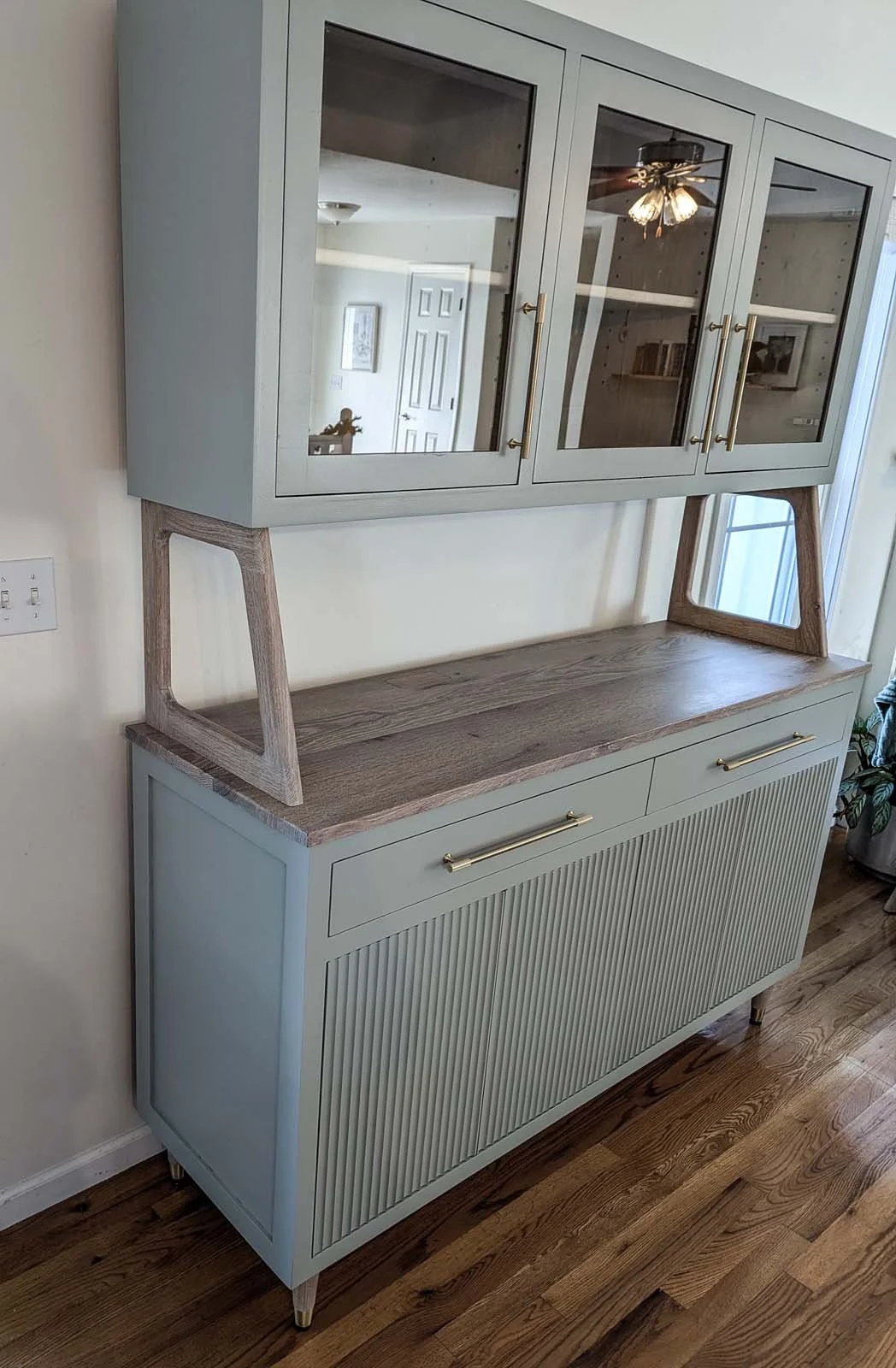 A modern sideboard with a wood top, light gray cabinet doors, and gold handles, topped by a glass-front cabinet, standing on four gold-tipped legs against a beige wall, with hardwood flooring underneath.
