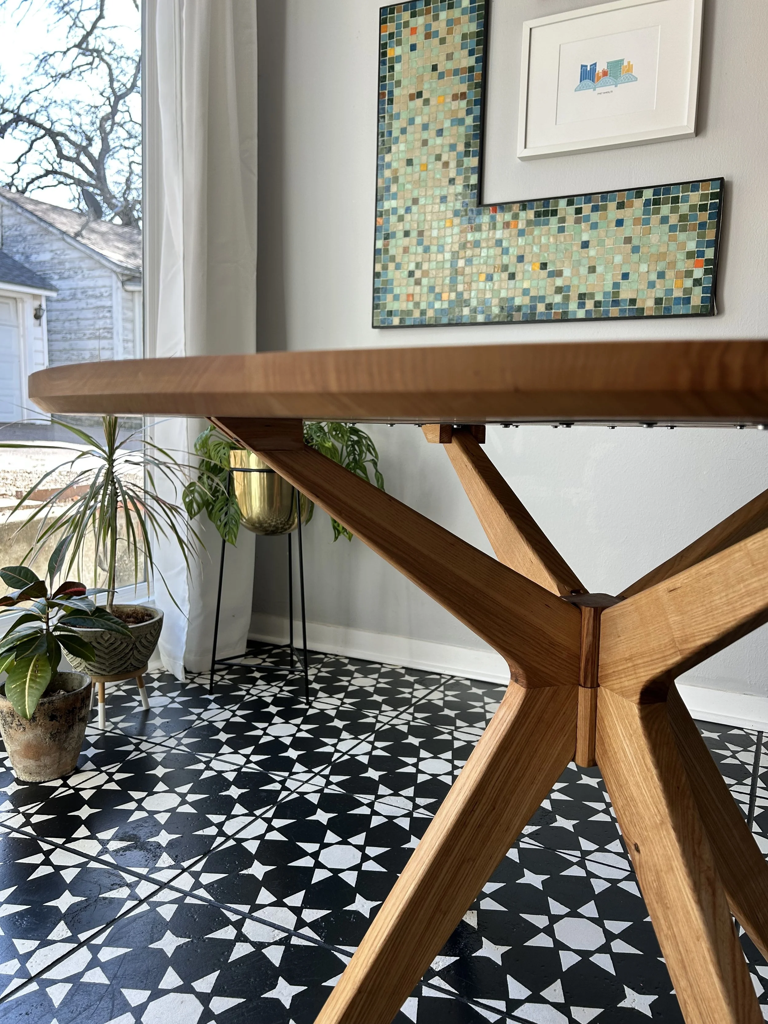 Close-up of a wooden table leg with a room in the background featuring artwork on the wall, patterned black and white floor tiles, and potted plants near a window.