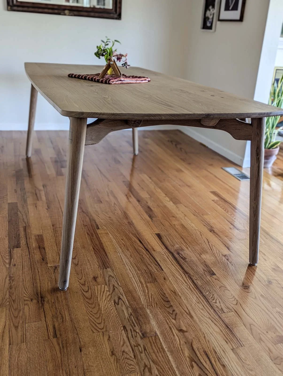 A wooden dining table with a small potted plant and a cloth on top, situated on a hardwood floor in a room with framed pictures on the wall.