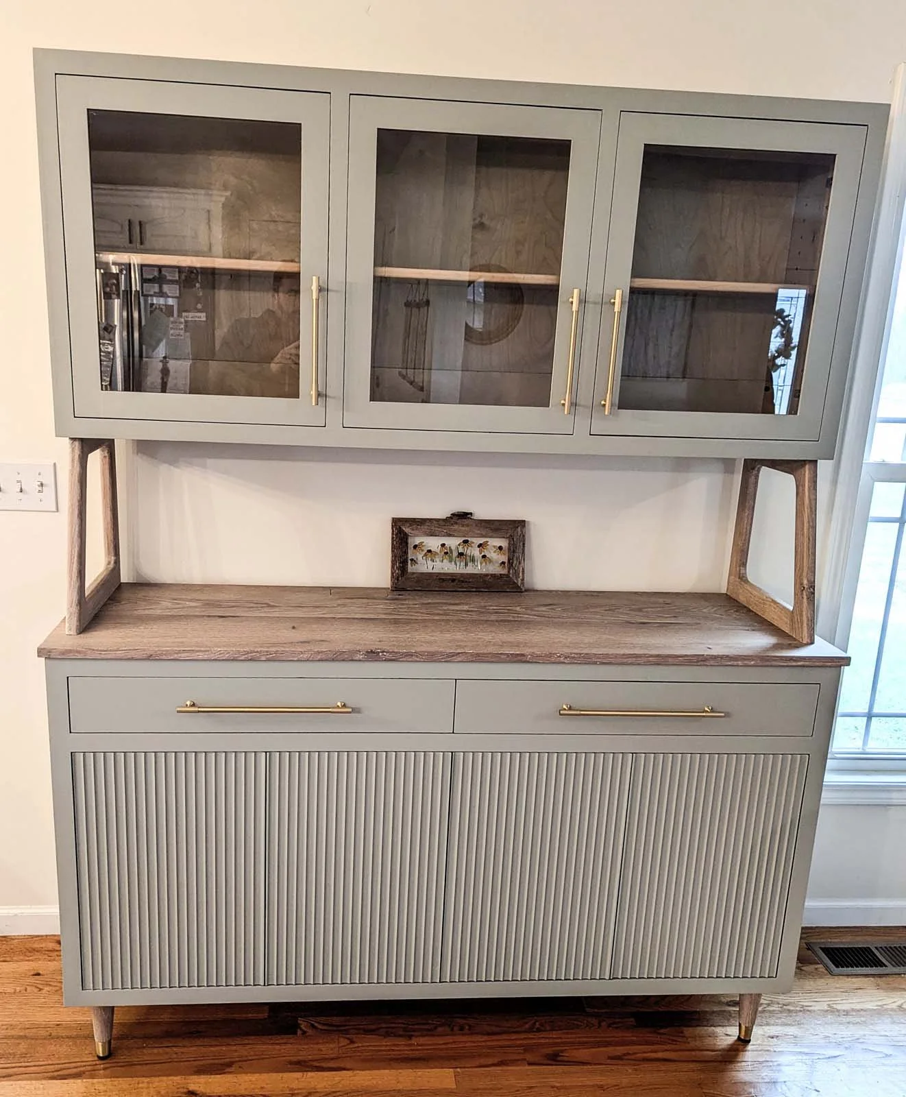 A light green cabinet with glass doors on the top section, a wooden countertop in the middle, and ribbed doors on the bottom, with gold handles and legs, against a white wall.