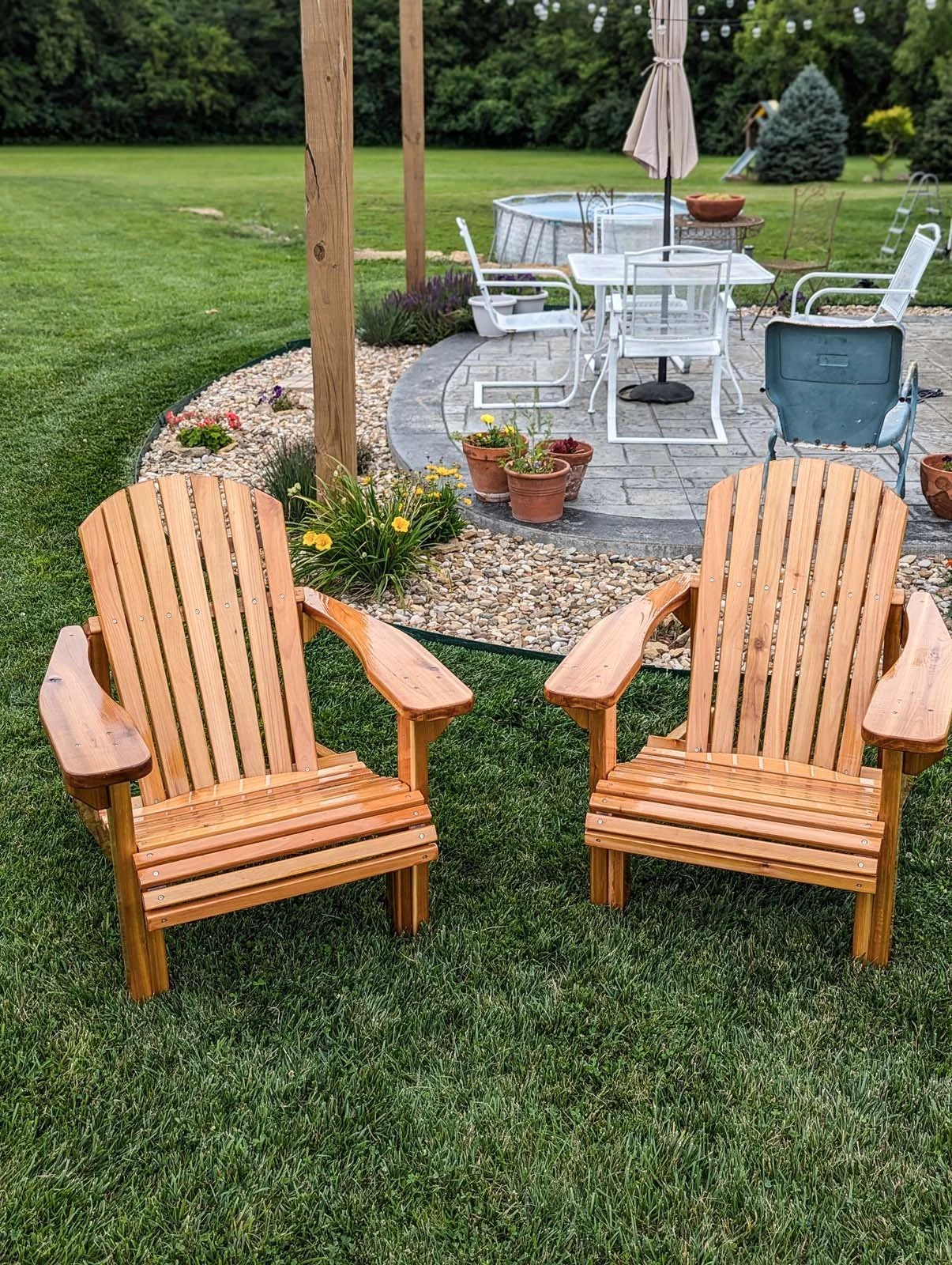 Two wooden Adirondack chairs on green grass facing a patio with outdoor furniture and potted flowers.