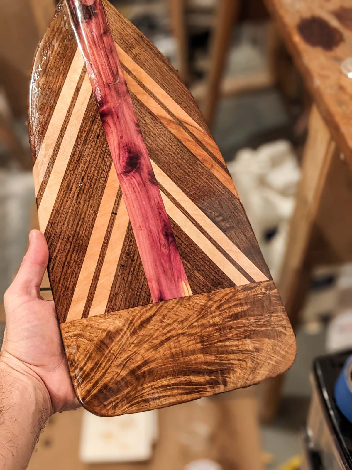 A hand holding a wooden cutting board with a geometric inlay pattern in various wood tones, including a central pinkish-red strip and a rounded bottom edge.