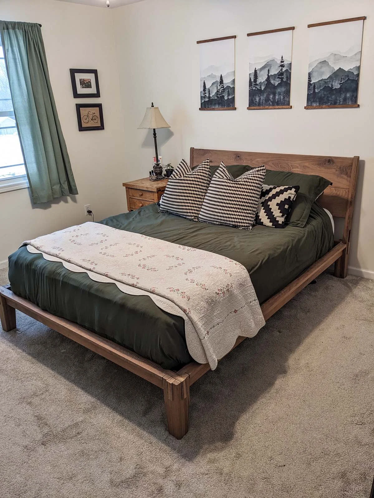 A cozy bedroom with a wooden bed frame, dark green bedding, and patterned pillows. A nightstand with a lamp and decor, framed pictures on the wall, and a window with green curtains.