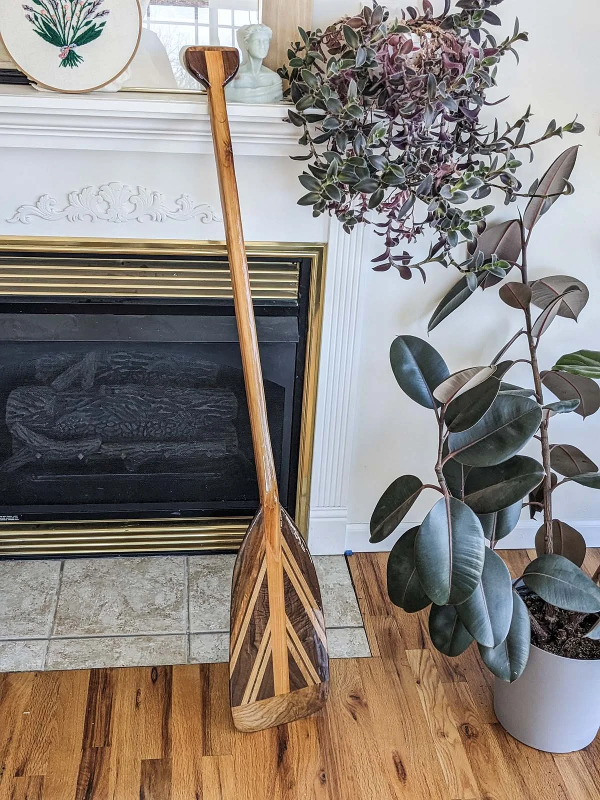 A wooden paddle with a chevron pattern stands upright against a fireplace. A potted plant with large, dark green and purple leaves is beside it, and decorative items, including a bust sculpture and framed art, are on the mantel.