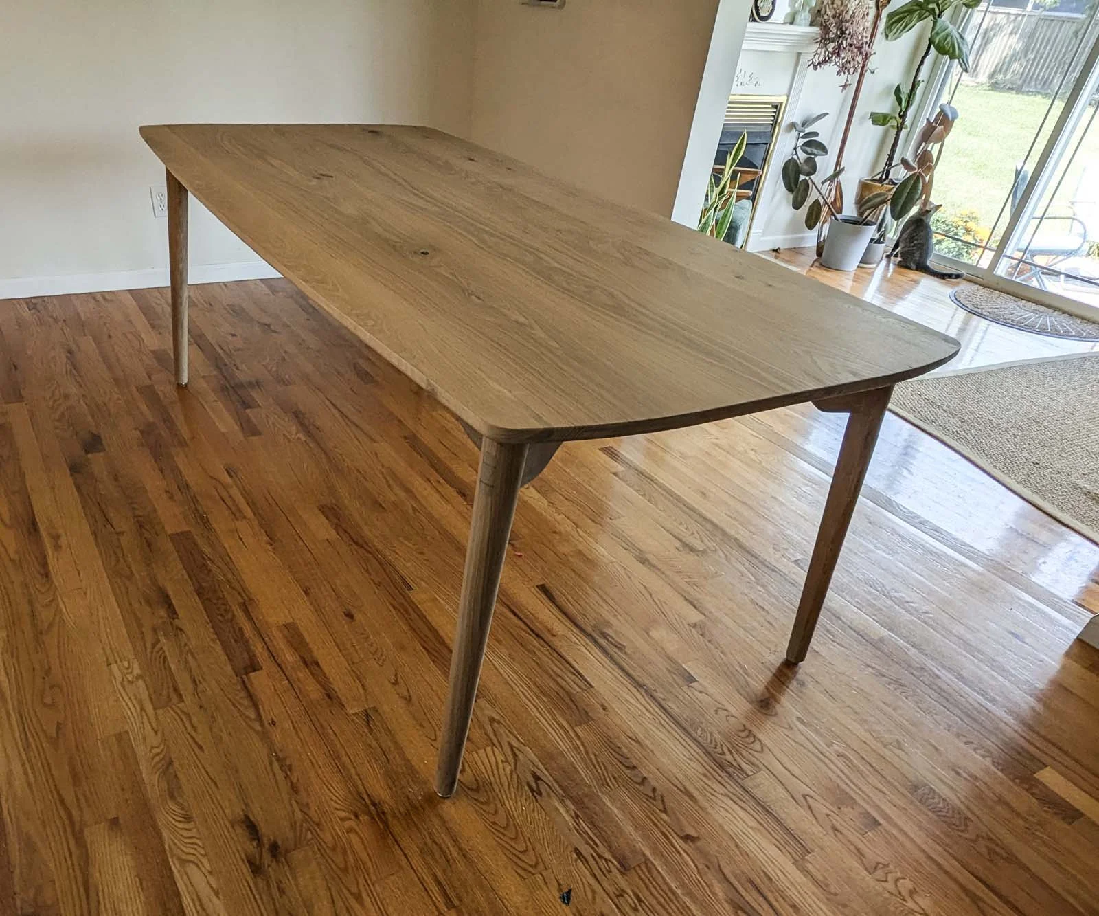 A wooden dining table with a smooth, natural finish in a room with hardwood flooring, near a sliding glass door and decorated with potted plants.