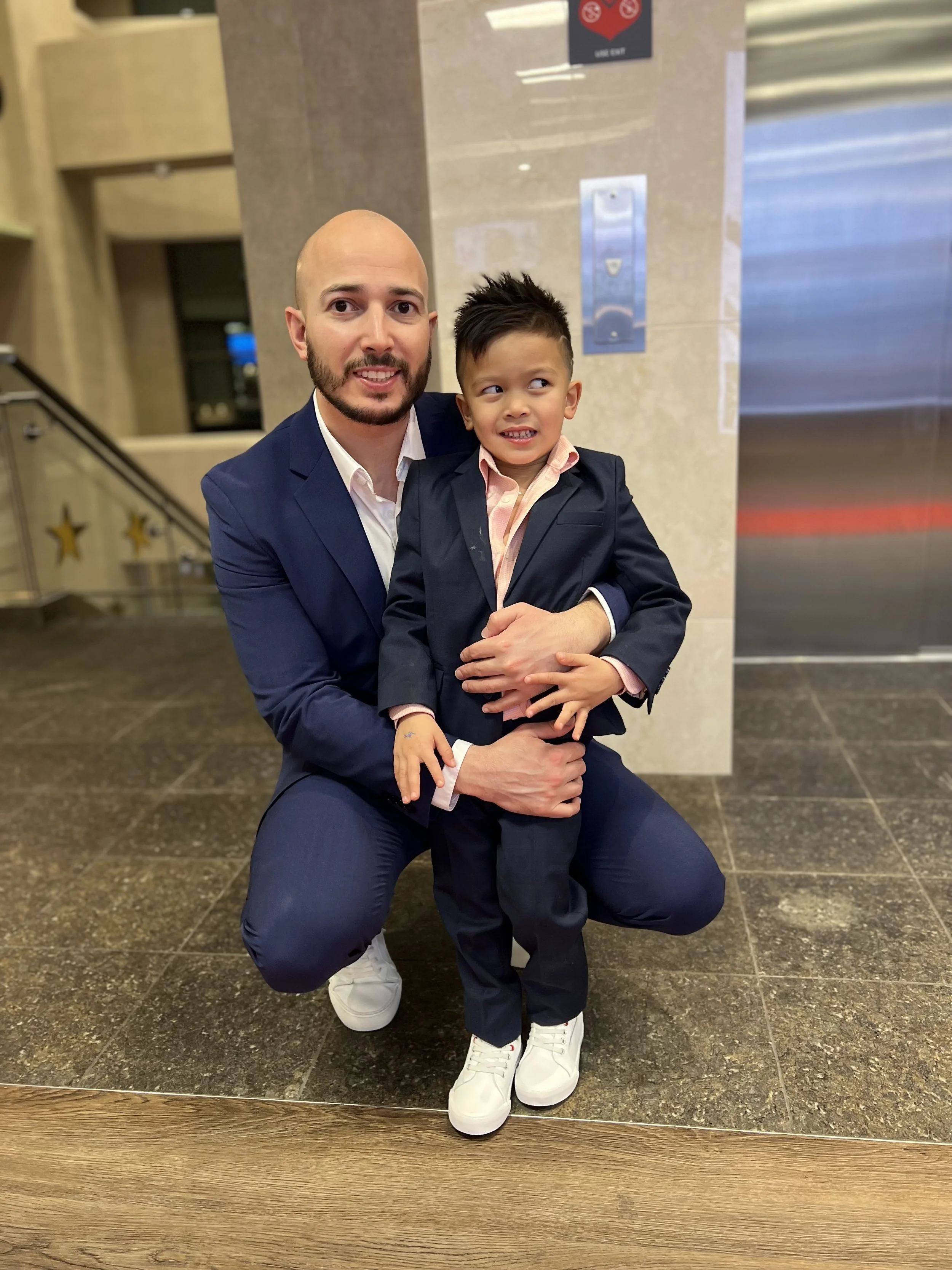 A man and a young boy dressed in suits pose together indoors. The man is kneeling with a smile, holding the boy, who appears a bit nervous or uncomfortable. The background shows an elevator and a wall with decorations, including some stars.
